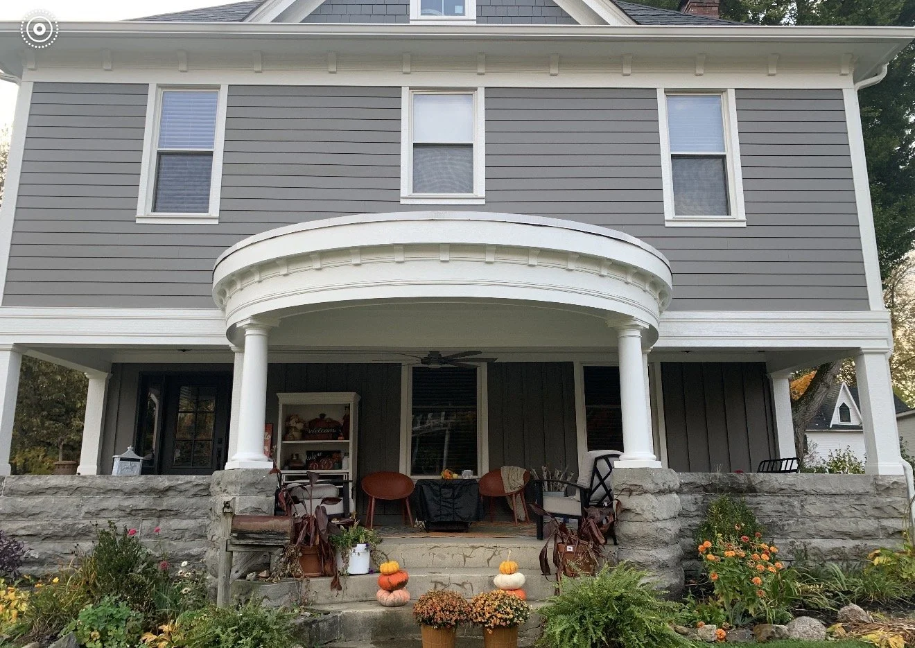 Front view of a two-story house with gray siding, white trim, and a curved porch supported by white columns. The porch is decorated with pumpkins and flowers, and there are outdoor chairs and a small table. The house has three windows visible on the second floor, and the garden in front features various plants and flowers.