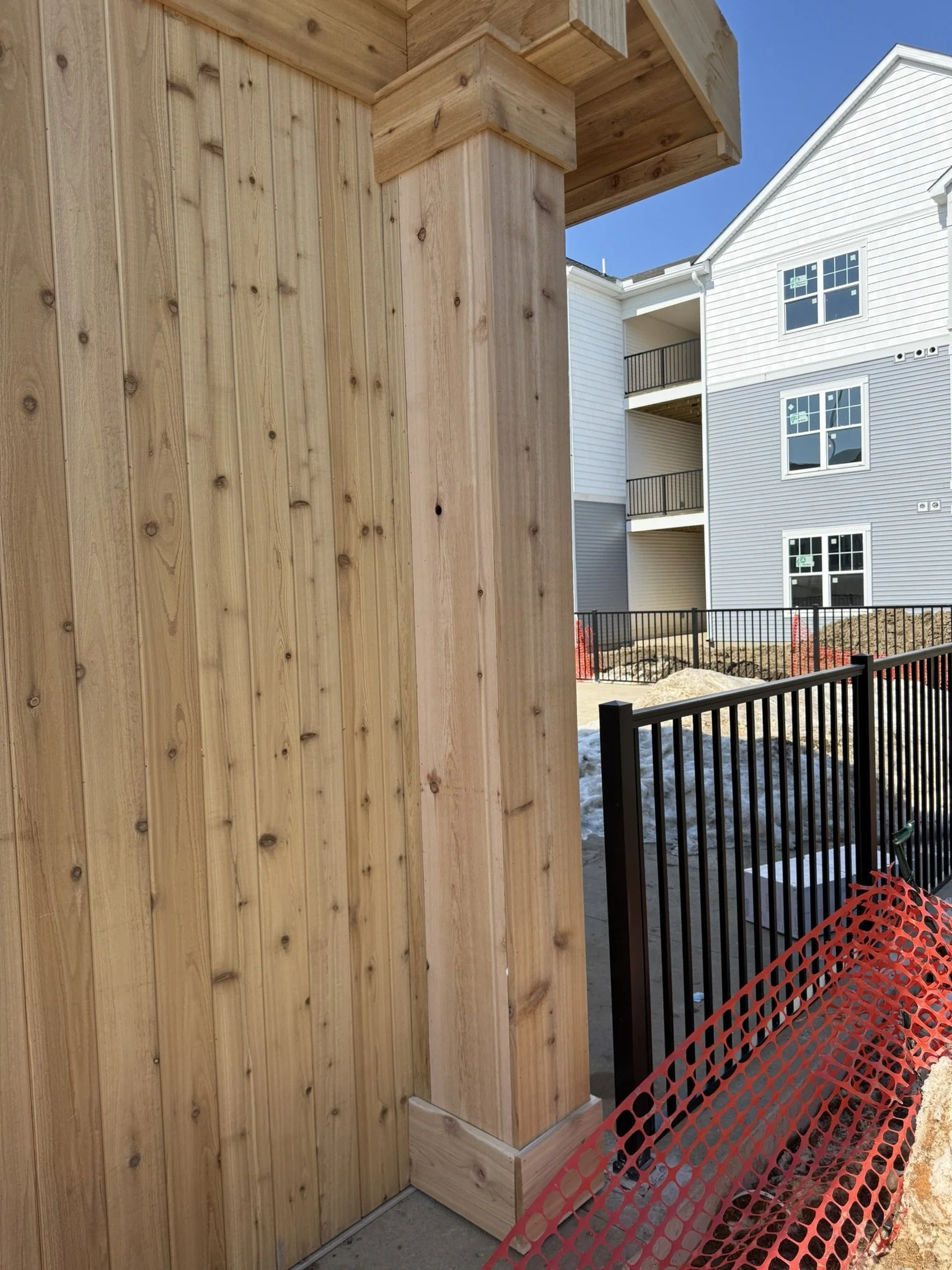 A wooden privacy fence with vertical planks and a decorative post cap. In the background, a white multi-story apartment building with black railings and several windows. Orange construction fencing is visible on the ground surrounding the area.