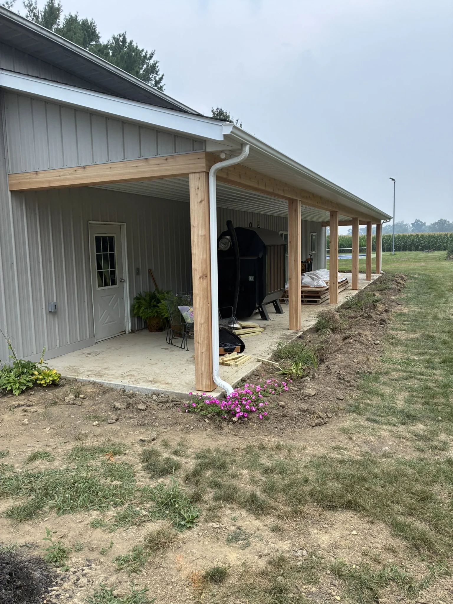A house with a metal exterior wall and a new wooden porch framework under construction. There is a door, garden plants, a pink flower, a grill, and pallets, with a field and trees in the background.