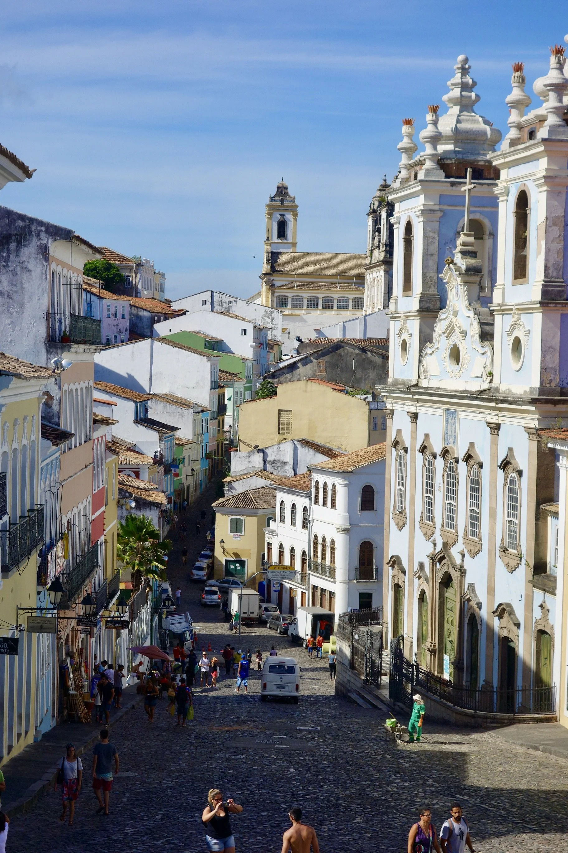 Street scene in Salvador da Bahia's historic Pelourinho neighborhood, with colorful buildings, a church with ornate white and blue architecture, and people walking on cobblestone streets under a blue sky.