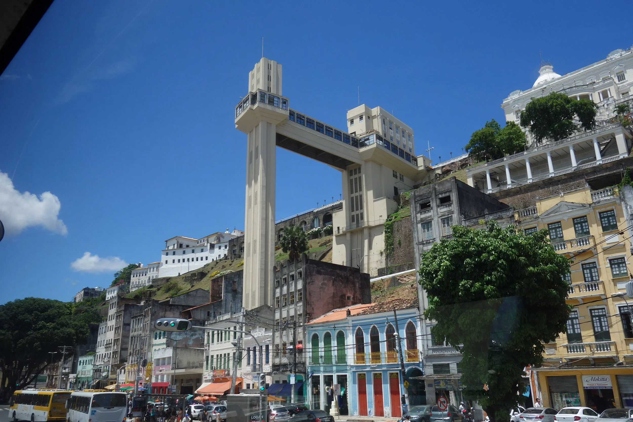 Street in Salvador da Bahia with colorful buildings, cars, buses, and people, with the famous "Lacerda" elevator on a hillside in the background, under a bright blue sky.