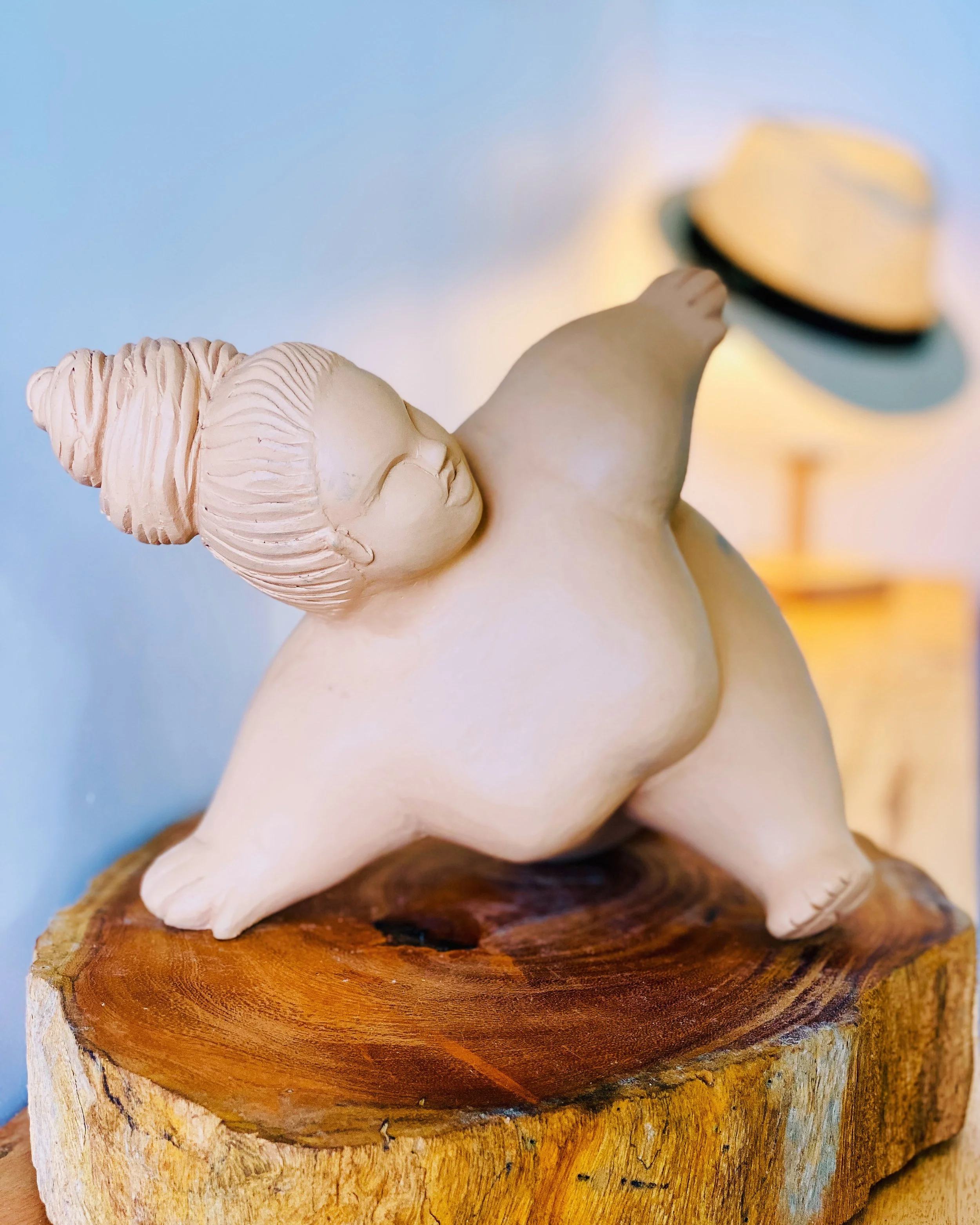 A ceramic sculpture of a woman on a wooden base, performing a capoeira move. In the background a lamp, covered with a straw hat with a black ribbon.