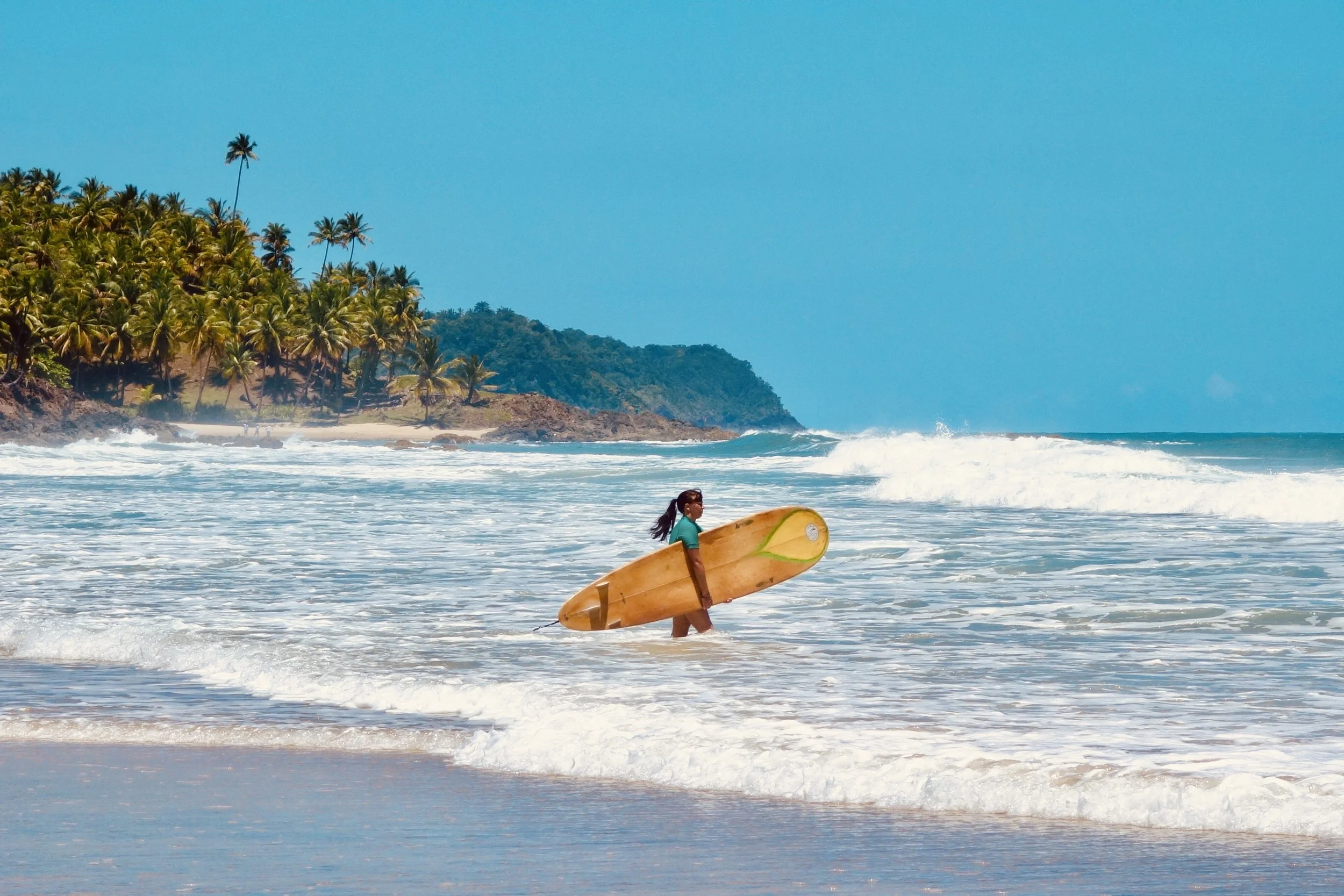 A woman holding a yellow surfboard while walking into the ocean waves at a beach in Itacaré with palm trees and a clear blue sky.