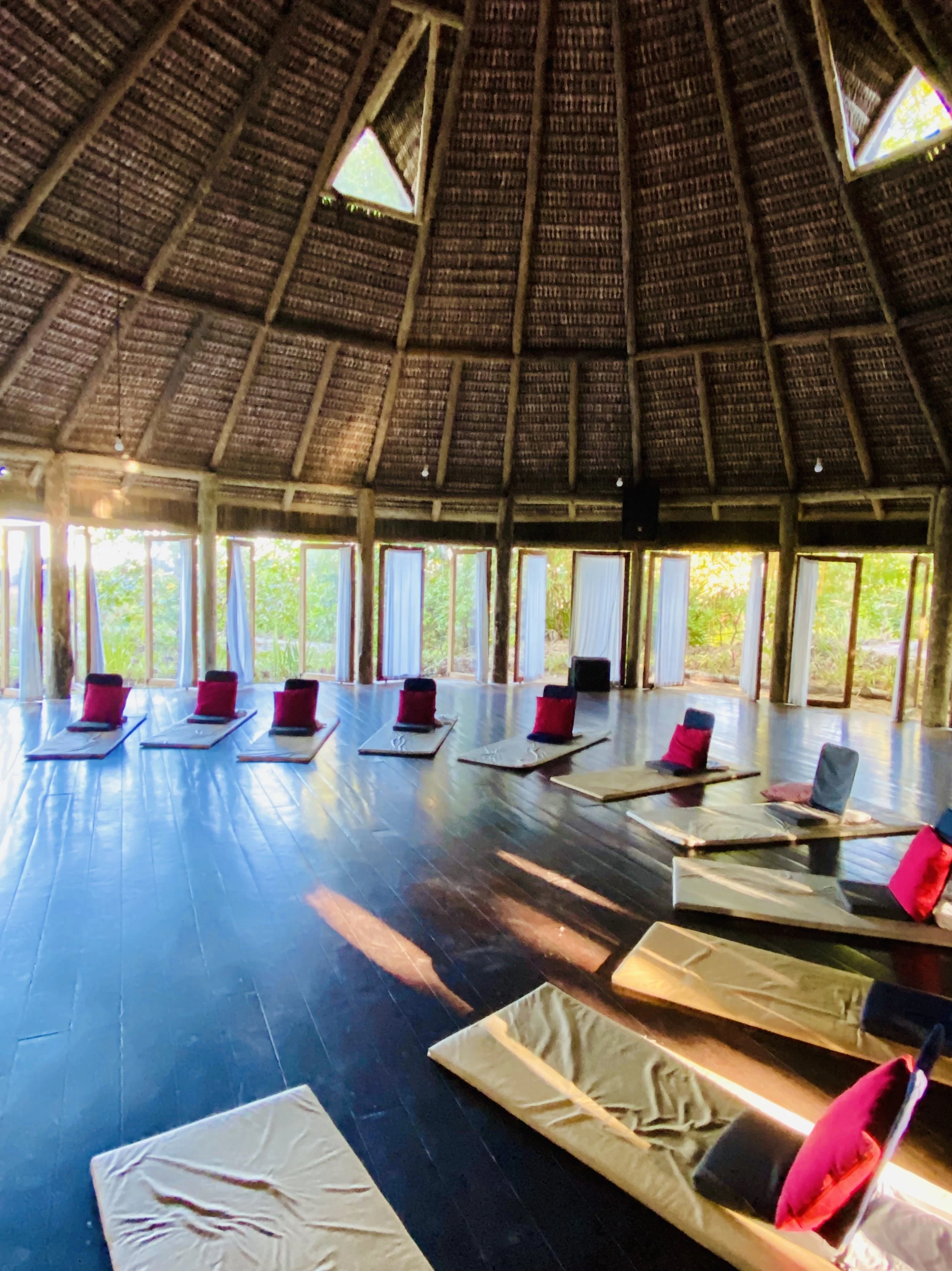 Interior of a circular, thatched-roof structure with windows letting in natural light. There are mats and cushions on the dark wooden floor, arranged in a semi-circle or line, likely for a meditation or yoga session.