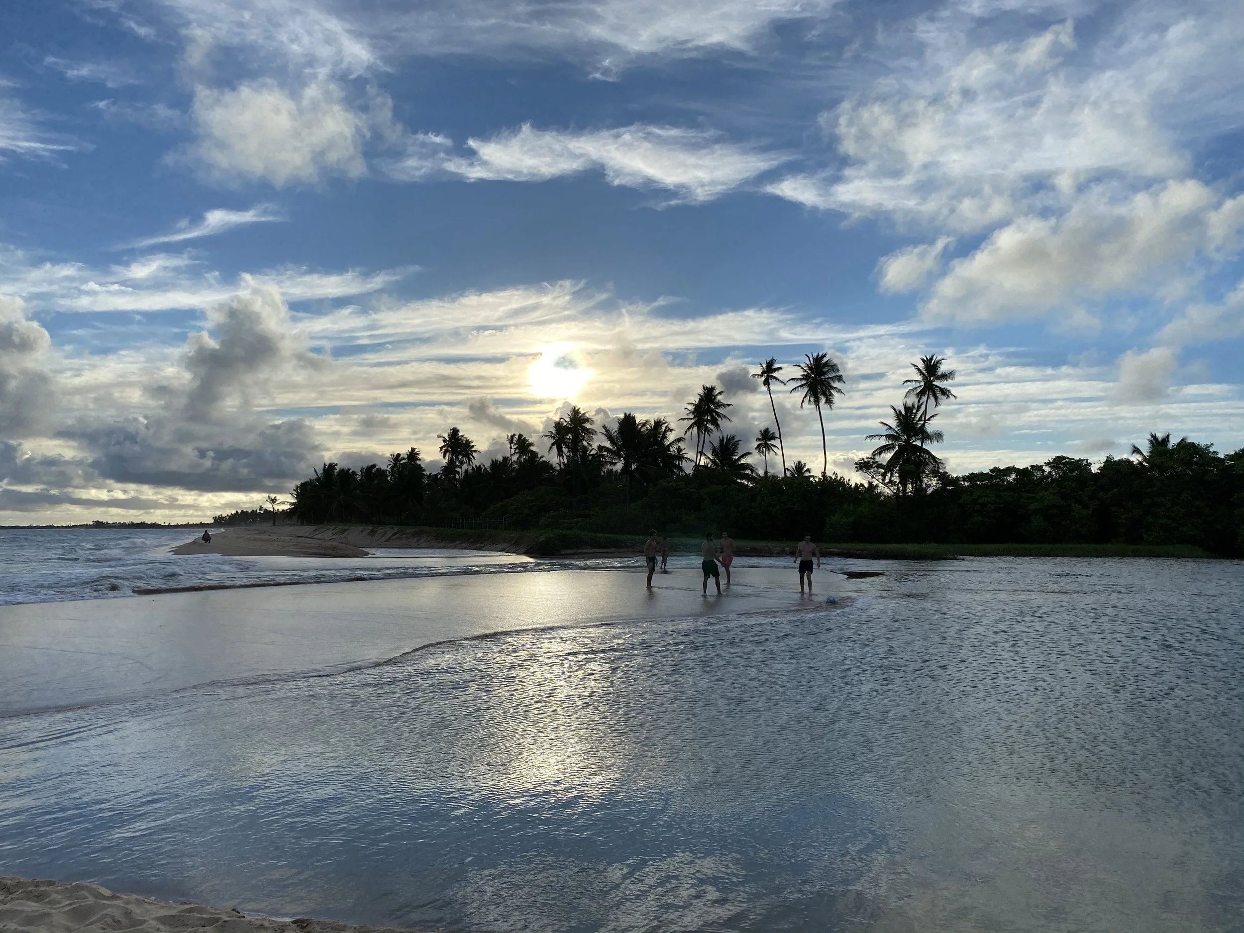A sandy beach covered by a shiny thin layer of seawather, shining in the evening light, several palm trees, and a partly cloudy sky with the sun low on the horizon. 