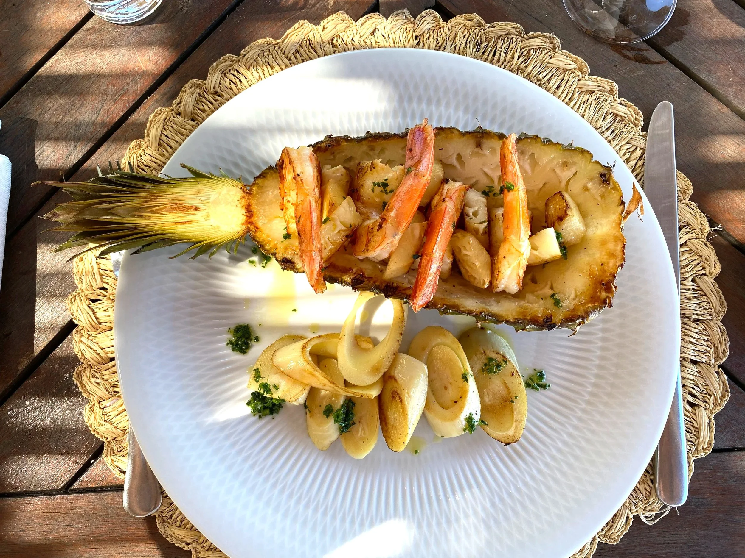 A sliced pineapple with shrimp and palm heart served on a white plate, placed on a woven placemat on a wooden table.