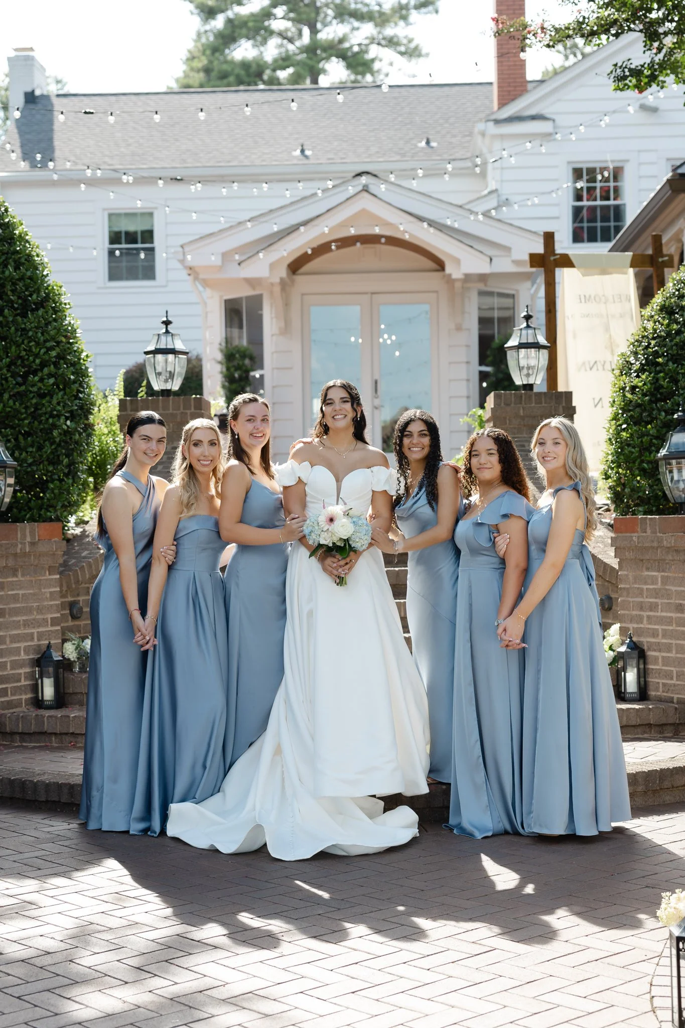 A bride in a white gown holding a bouquet and six bridesmaids in light blue dresses standing on steps outside a white house decorated with string lights.