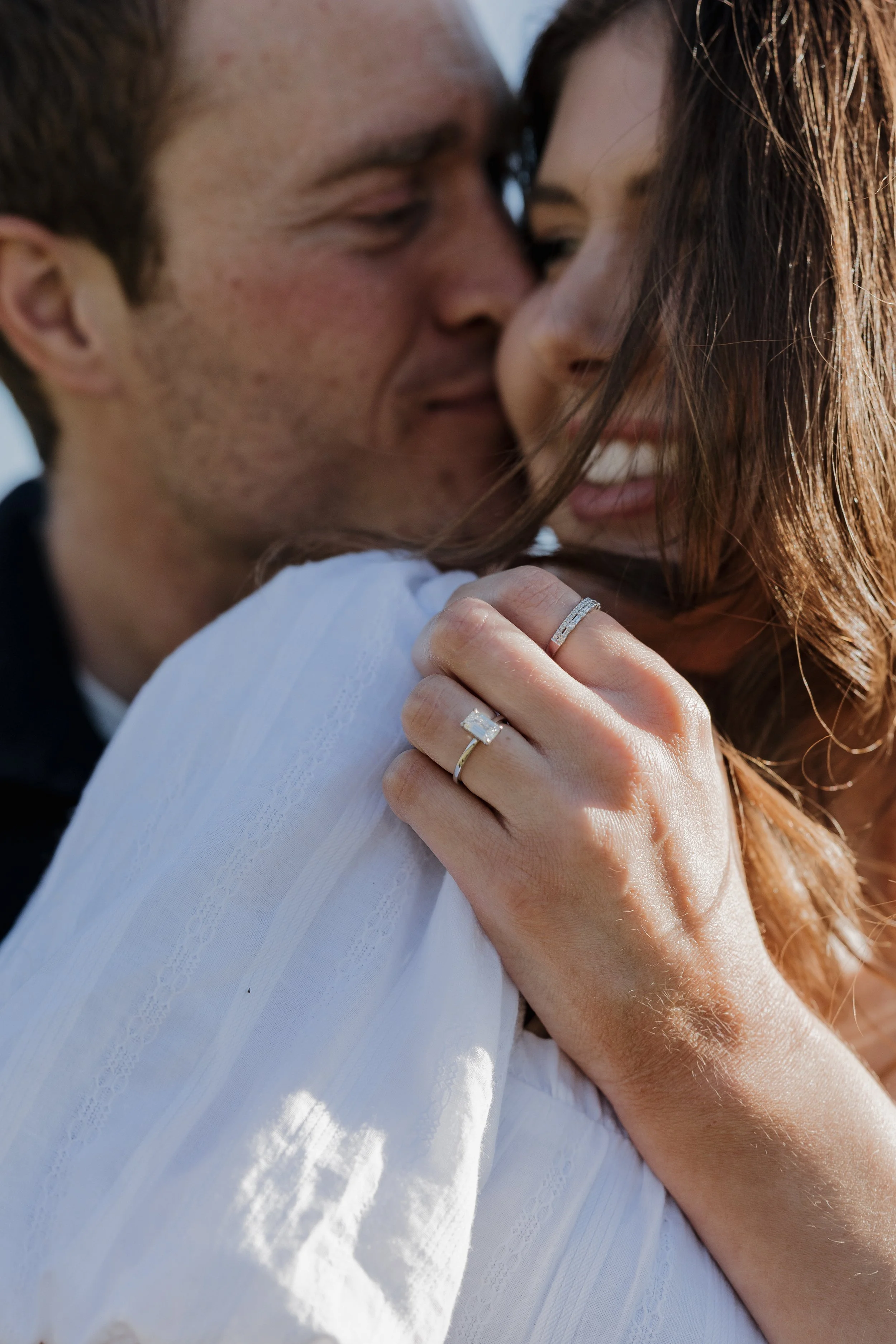 Close-up of a couple embracing. The man has light skin and short brown hair, smiling with eyes closed, and the woman has light skin with long brown hair, smiling with visible teeth. They are wearing wedding rings, and the woman has her hand on the ma