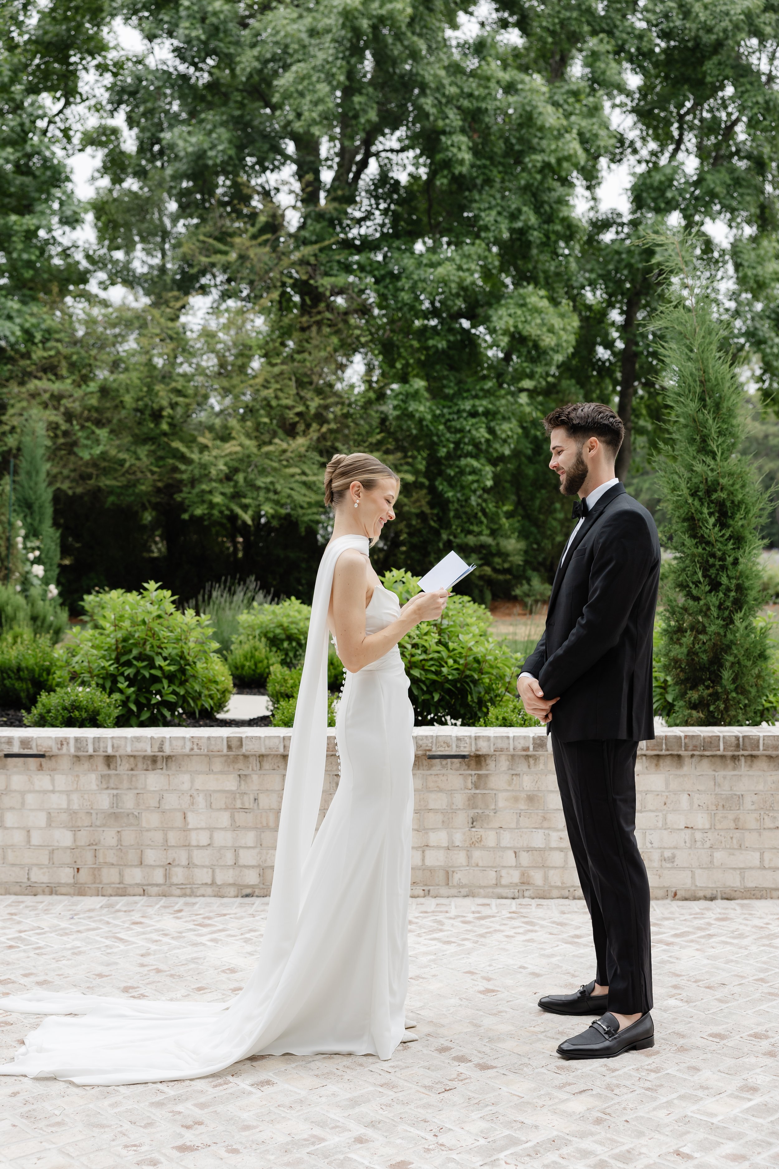 Bride and groom exchanging vows outdoors, with the bride reading from a paper and the groom holding hands, surrounded by green trees and shrubs.