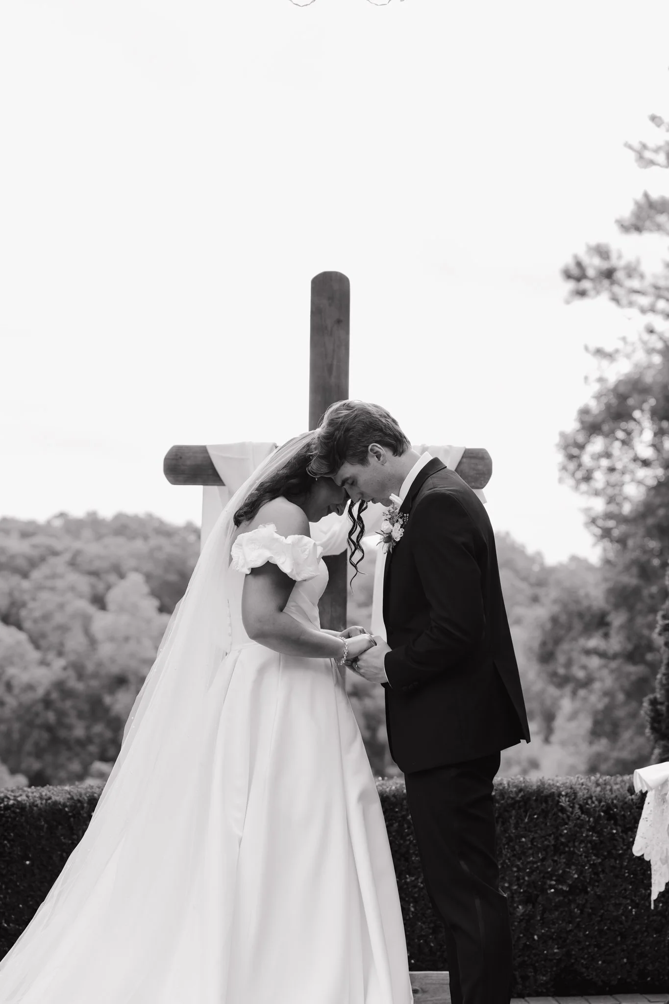 A bride and groom leaning head to head in front of a cross during a wedding ceremony outdoors.