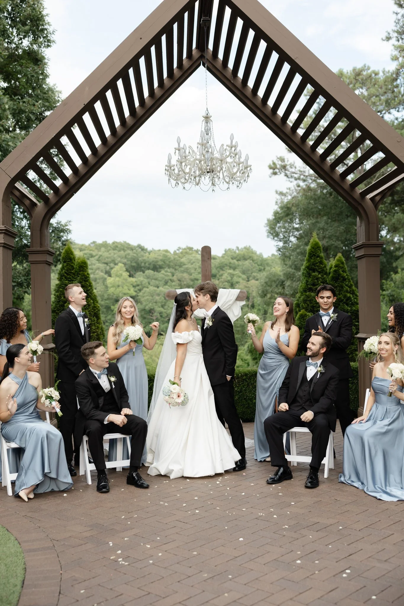 A wedding ceremony outdoors with a couple kissing in the center, surrounded by bridesmaids and groomsmen in formal attire, under a pavilion with a chandelier, with trees and a landscape in the background.