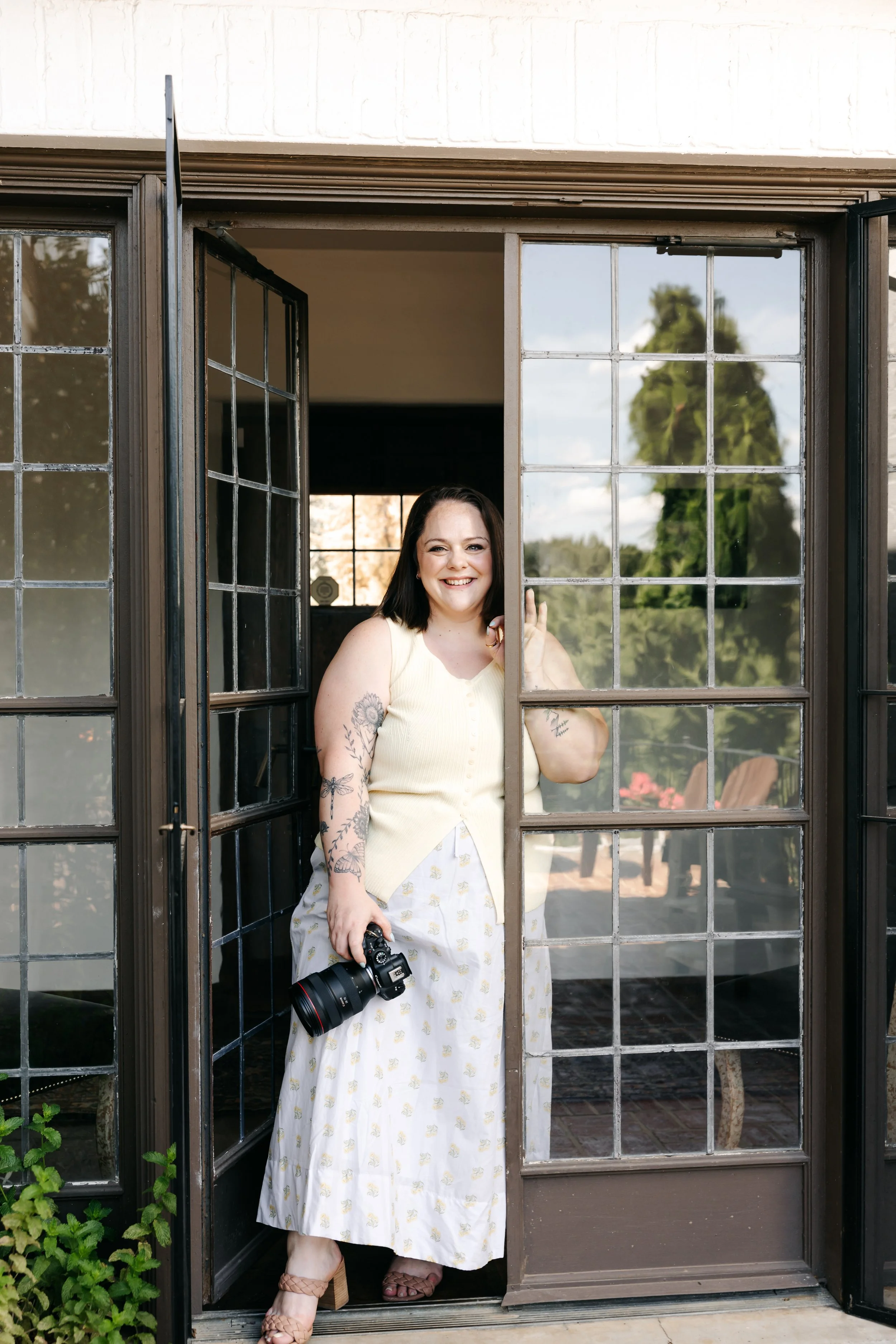A woman stands inside a large window opening, smiling and holding a camera with her left hand. She has shoulder-length dark hair, tattoos on her arms, and is wearing a light yellow sleeveless top and a white skirt with a yellow pattern. She is outdoors with trees and a patio visible in the background.