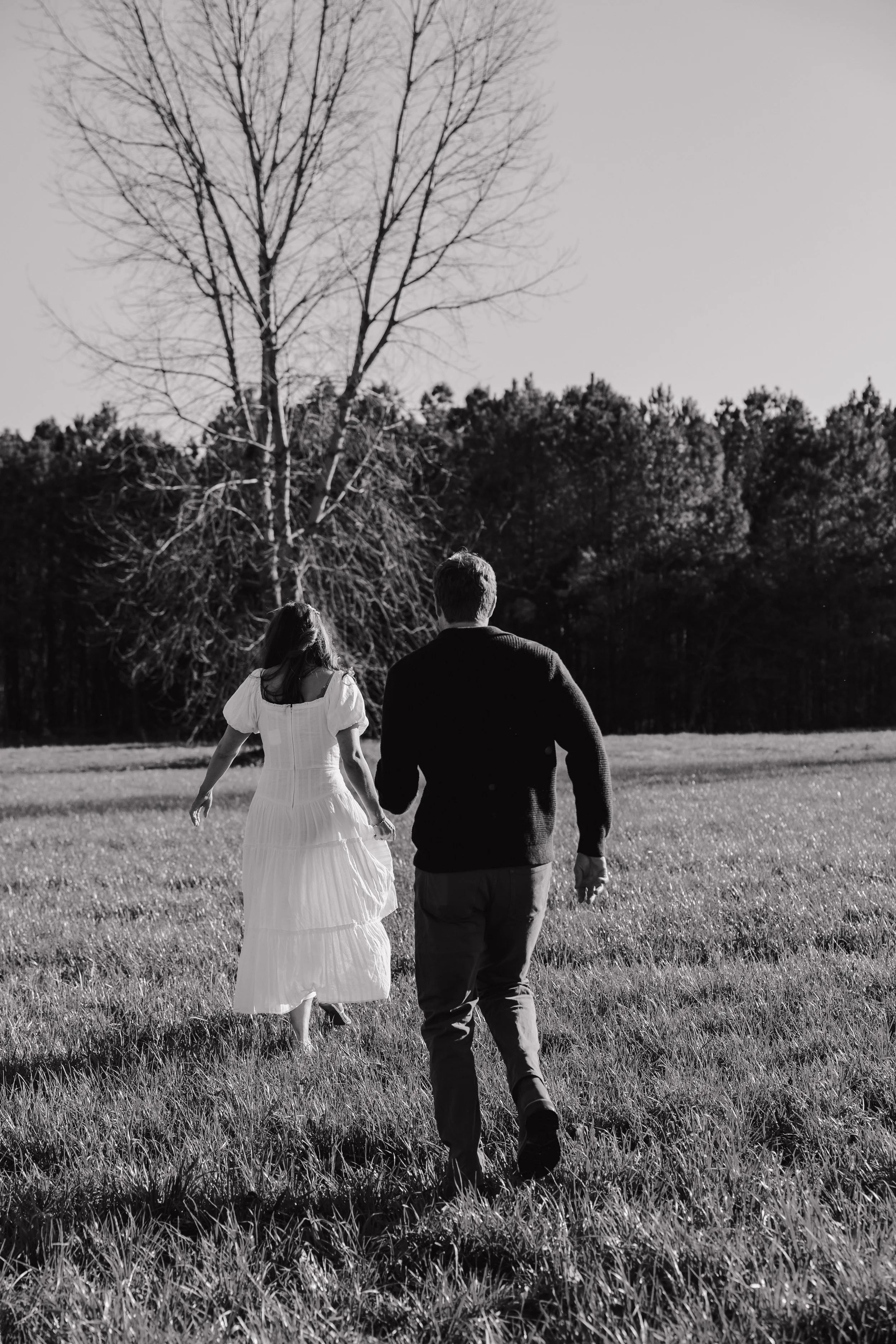 A black and white photograph of a couple walking hand in hand through a grassy field, with trees in the background and a clear sky above.