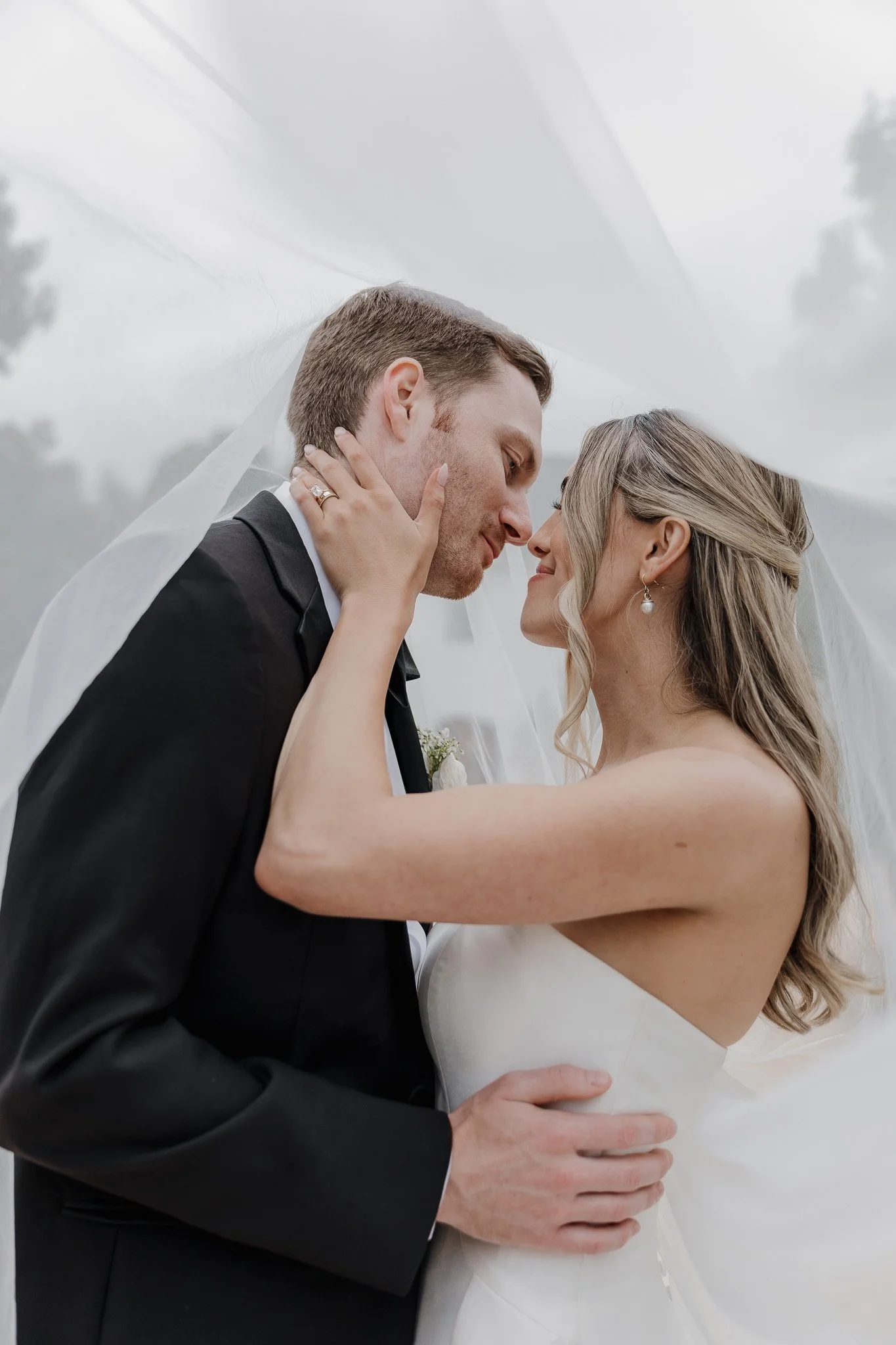 A newlywed couple gazing into each other's eyes, with the bride touching the groom's face and holding his shoulder, both dressed in wedding attire, under a white veil outdoors.