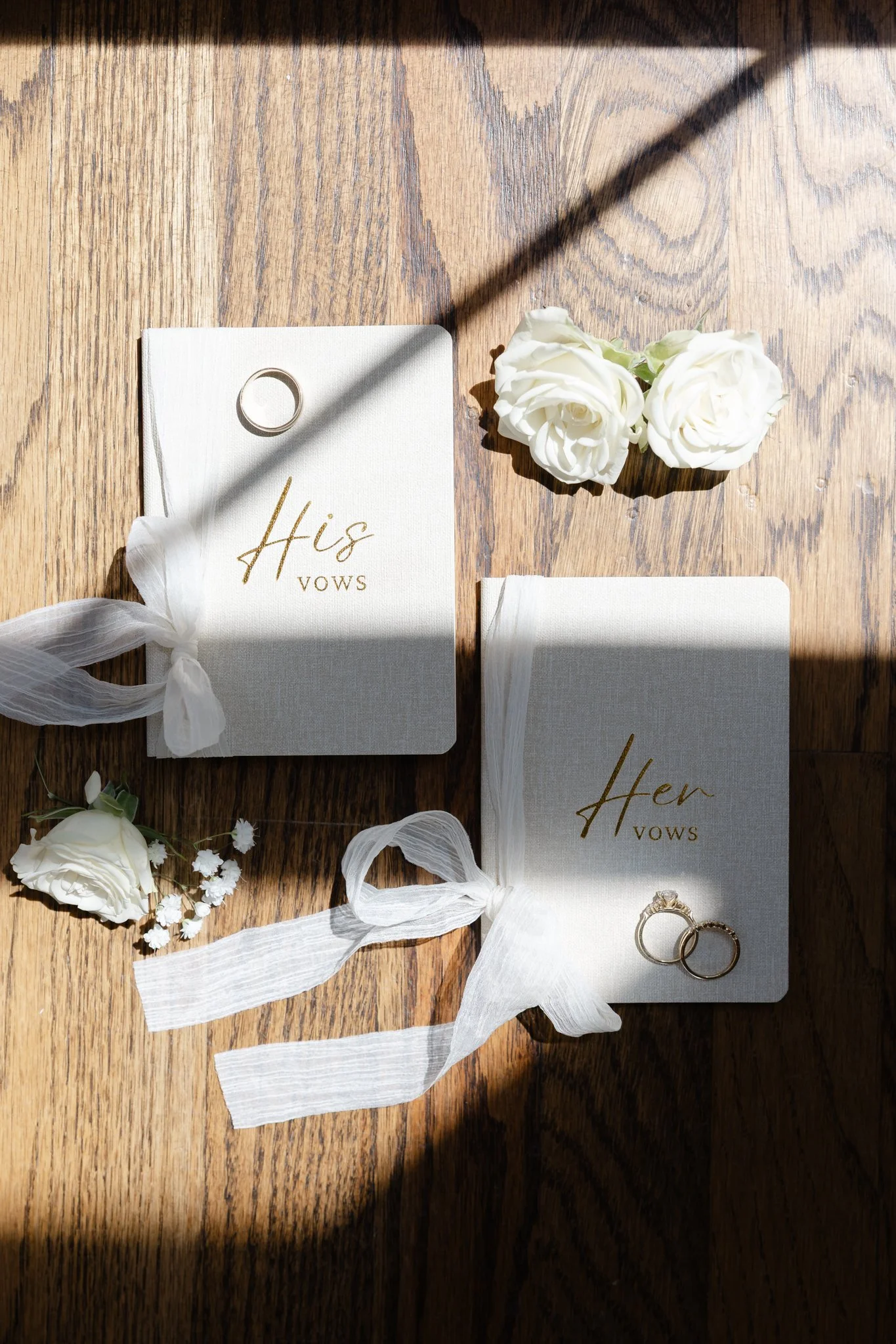 Wedding vows book labeled 'His Vows' and 'Her Vows' with wedding rings on top, white roses, and small white flowers on a wooden surface, illuminated by sunlight.