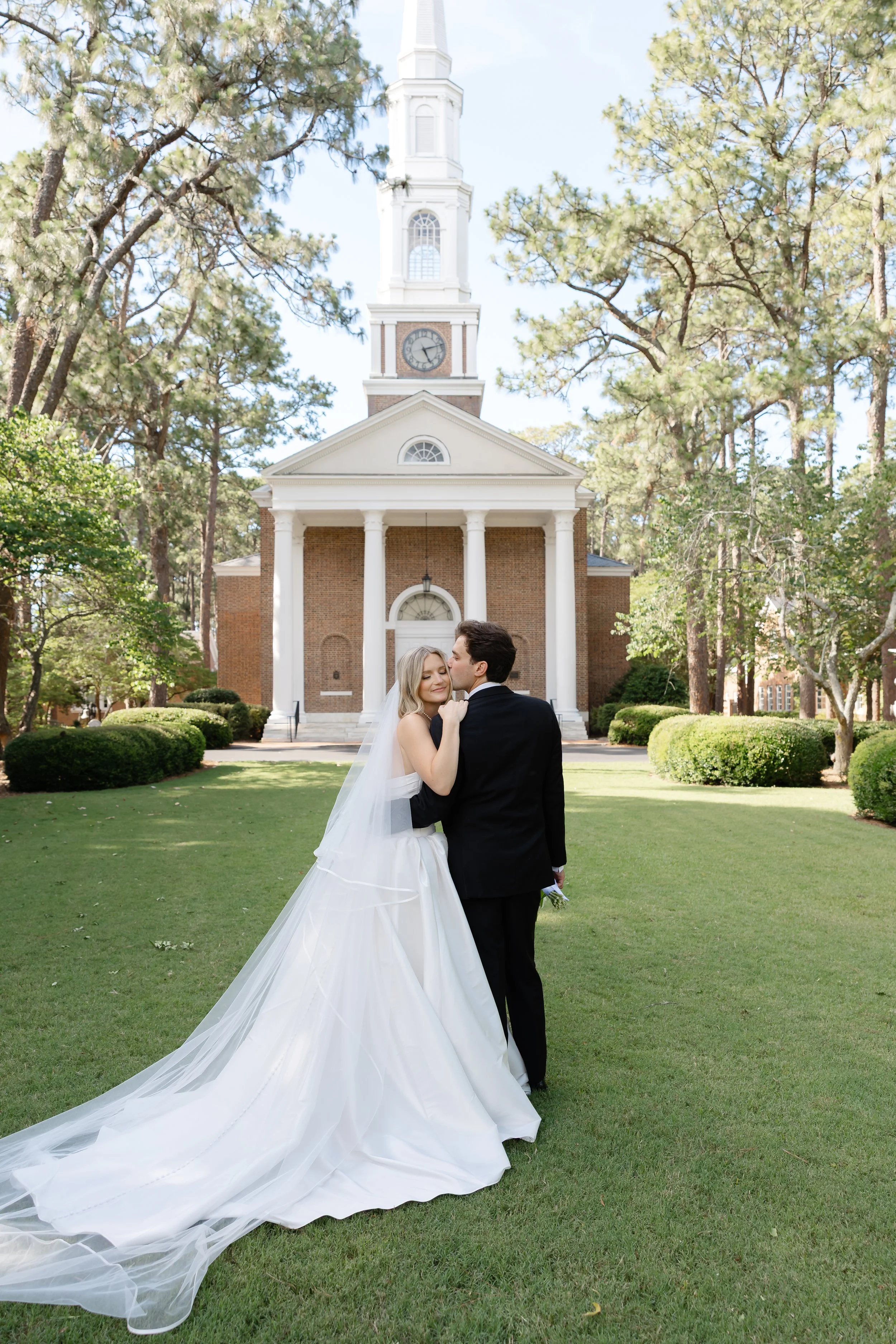 A bride and groom standing on a lawn in front of a white church with columns, trees, and a clock tower, sharing a kiss.