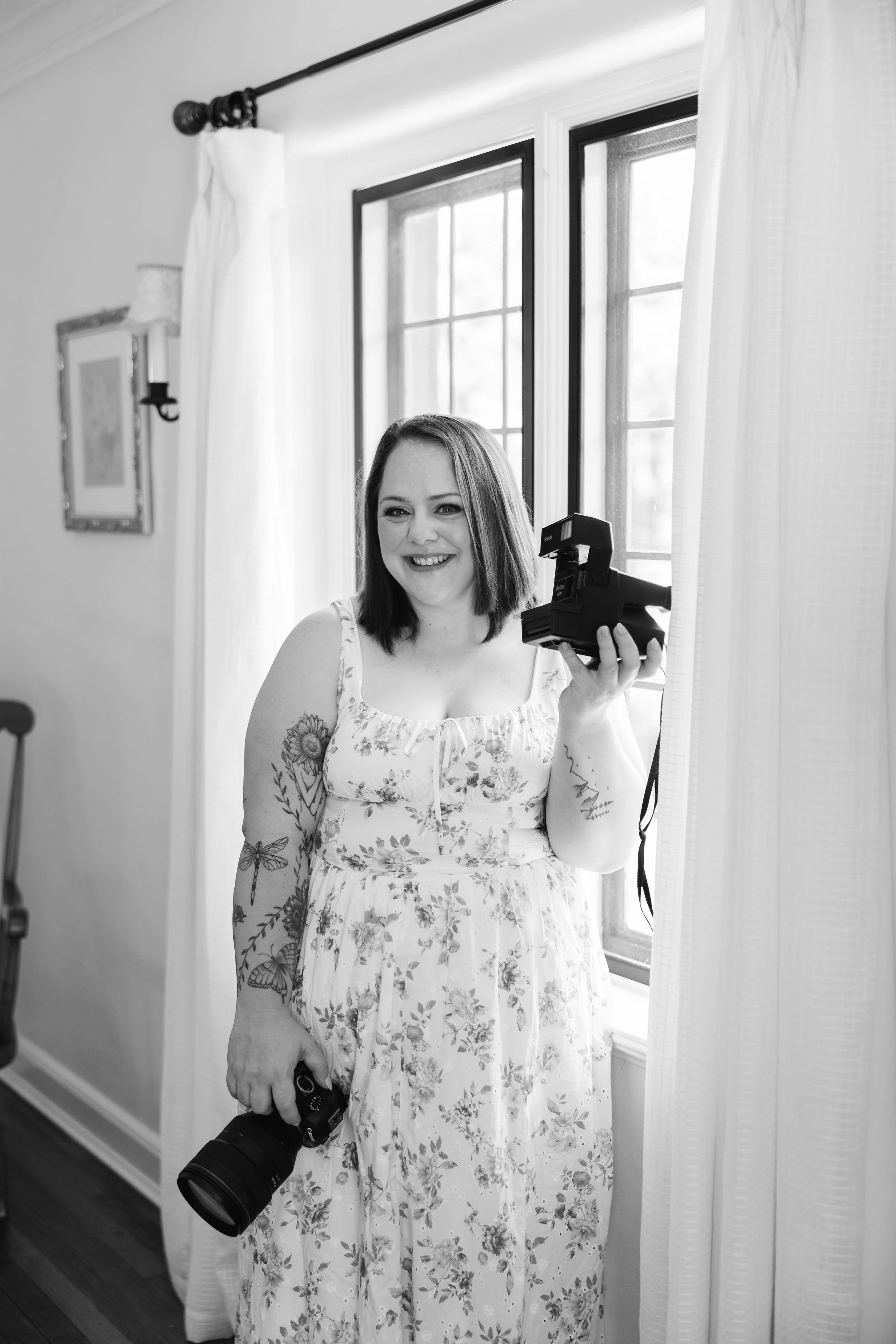 A woman with shoulder-length hair taking a selfie in front of a mirror, holding a camera in one hand and a phone in the other. She is wearing a floral dress and has tattoos on her arm, with a window and curtains in the background.