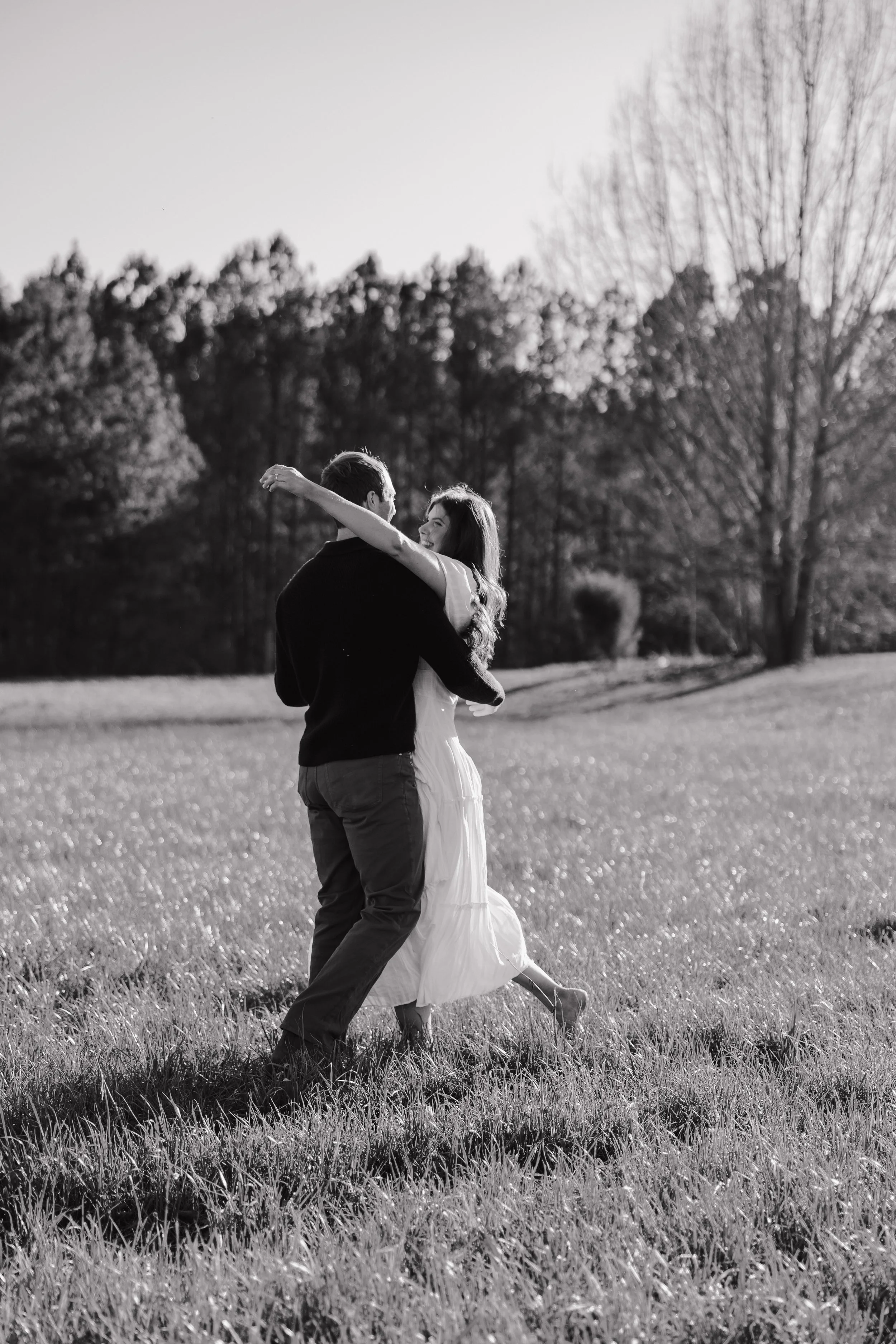 A black-and-white photo of a couple embracing and dancing in a grassy field outdoors during daytime, with trees in the background.