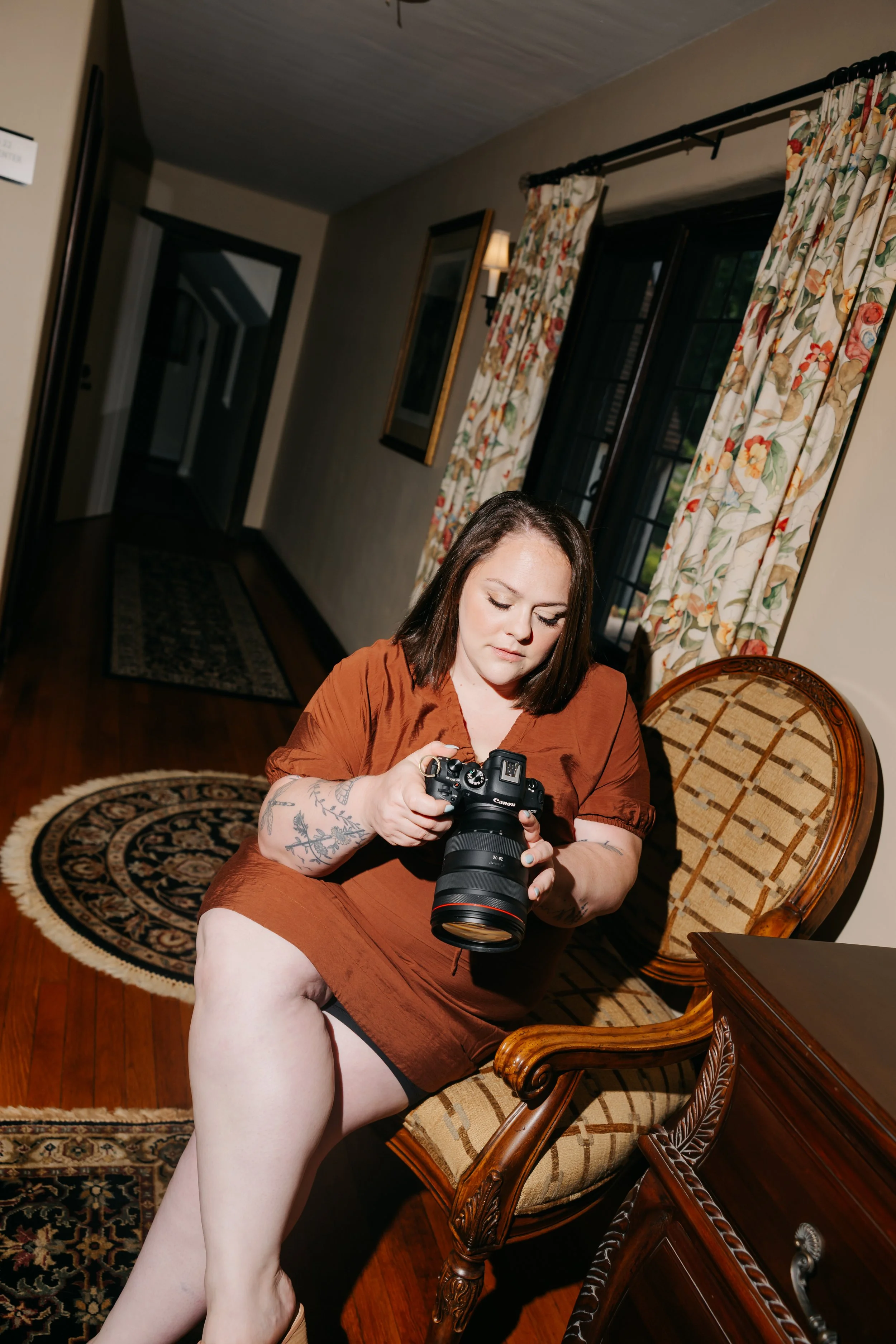 A woman with tattoos on her arm is sitting on a vintage upholstered chair inside a warmly lit room with floral curtains, holding a professional camera, with one leg raised and exposed.