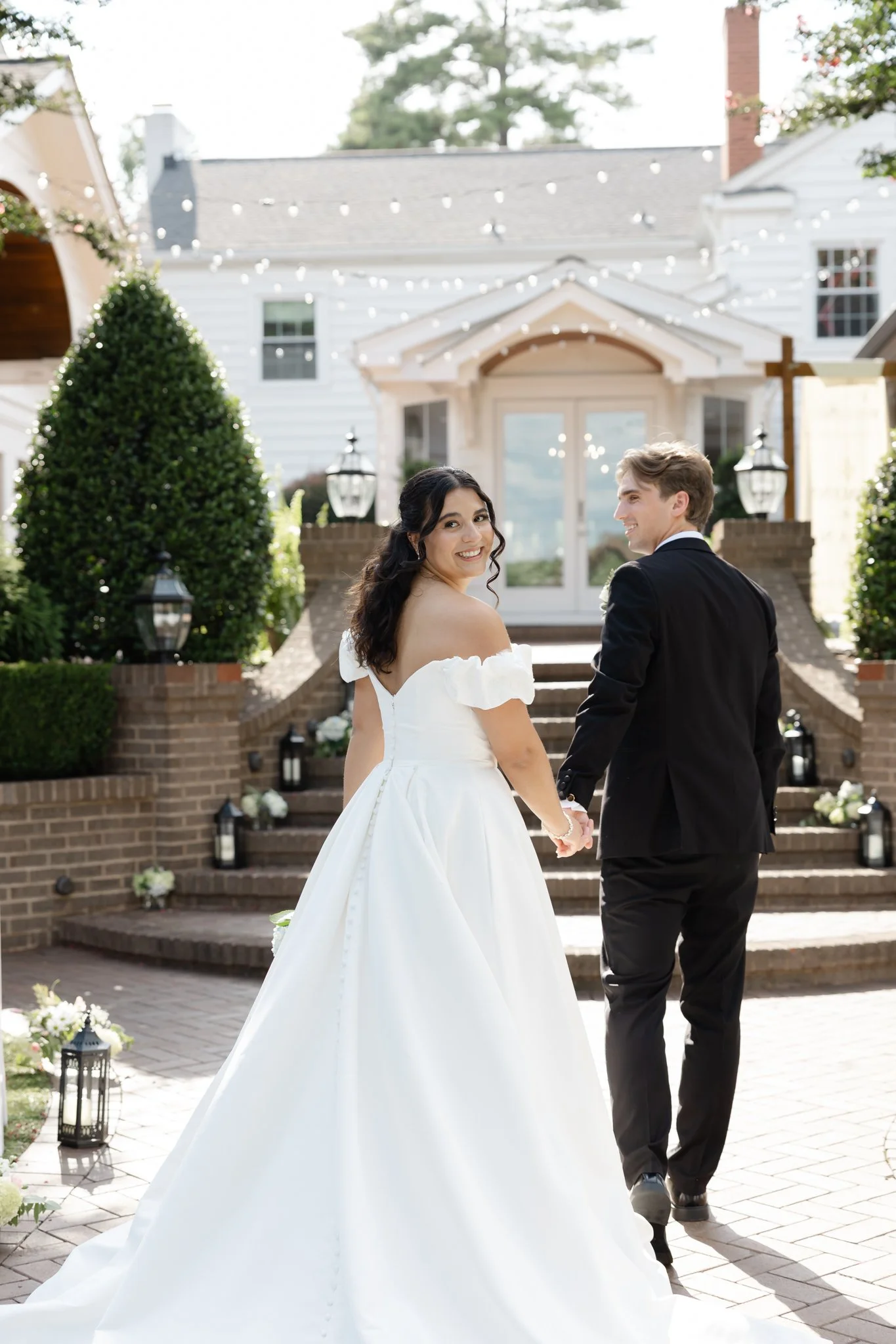 A bride and groom holding hands outside a white house with string lights and lanterns, smiling at each other on a sunny day.