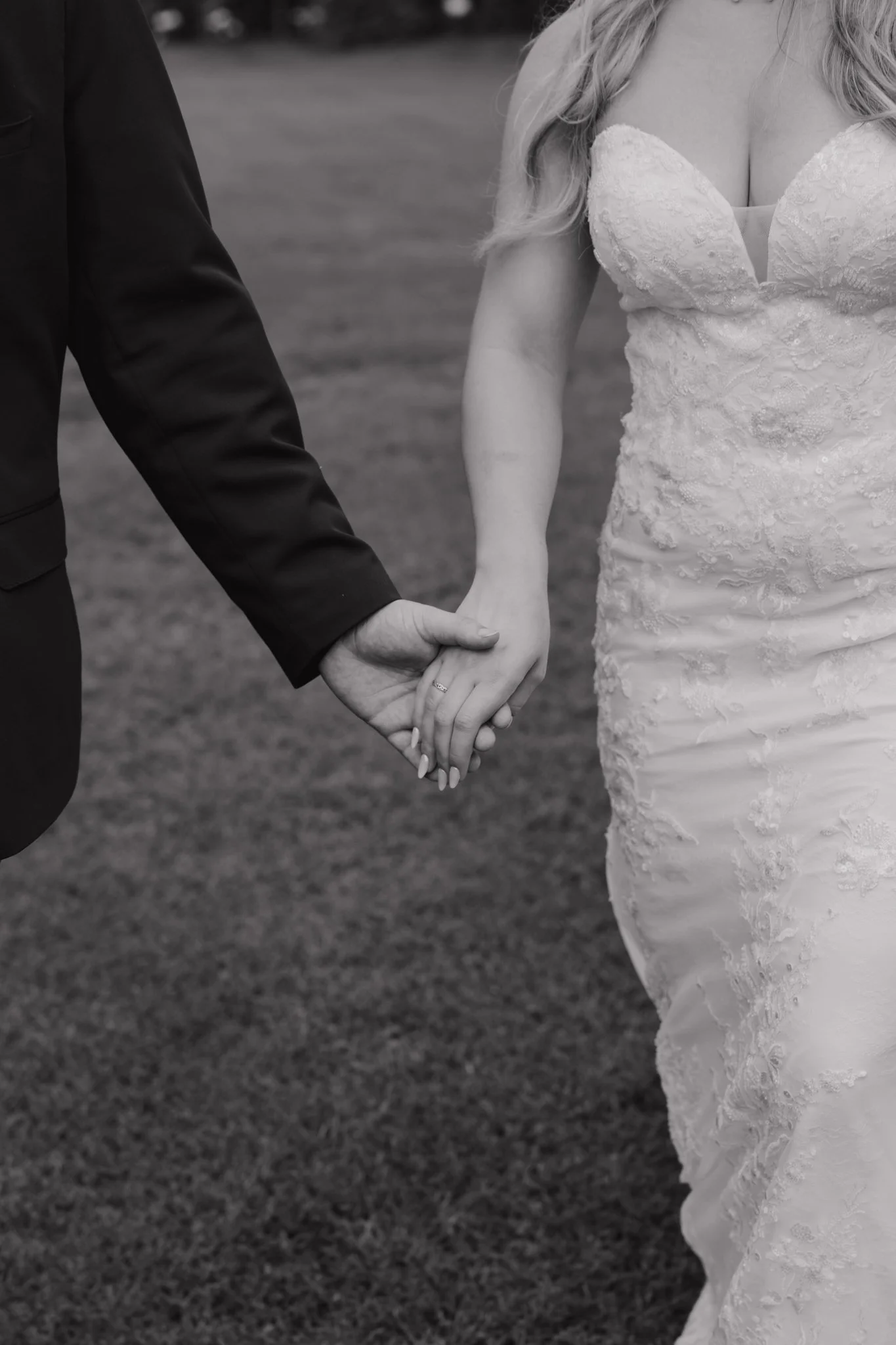 Close-up of a bride and groom holding hands outdoors, with the bride wearing a lace wedding dress and the groom in a dark suit, in black and white.