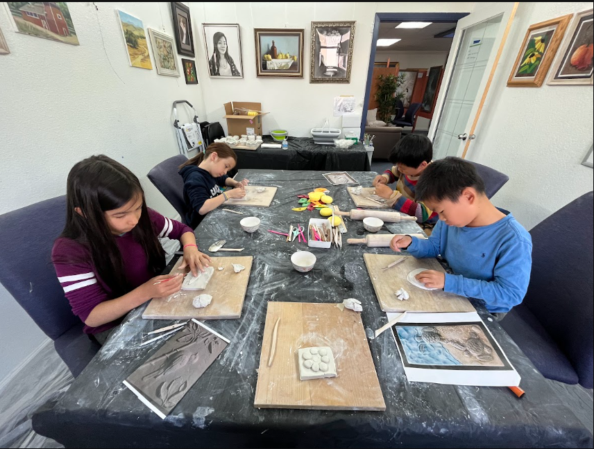 Four children sitting at a black table, working on art projects with clay and tools, in a room with artwork on the walls.