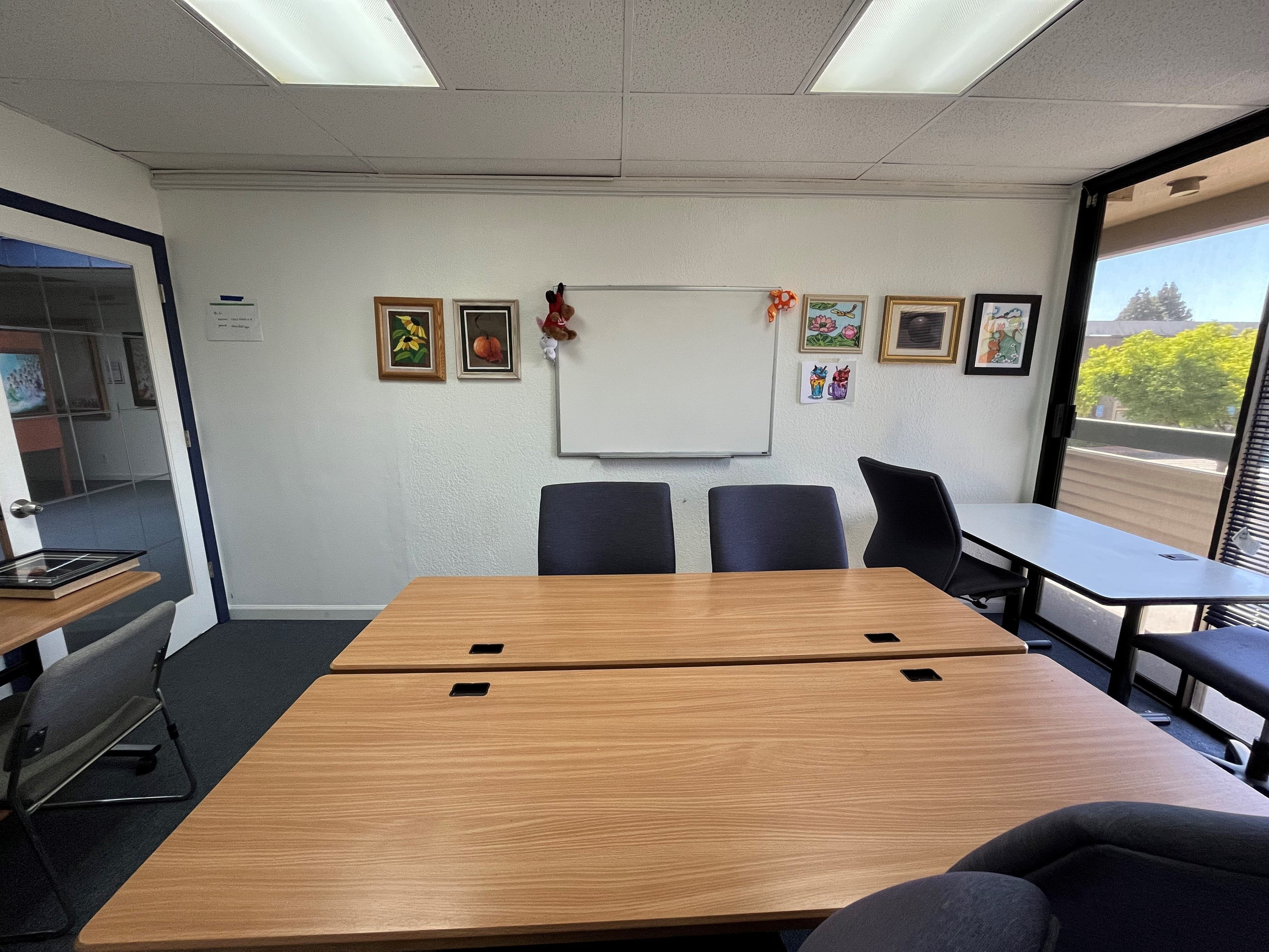 Inside of a conference room with a long wooden table, black chairs, framed artwork on white walls, a whiteboard, and large windows showing outside greenery.