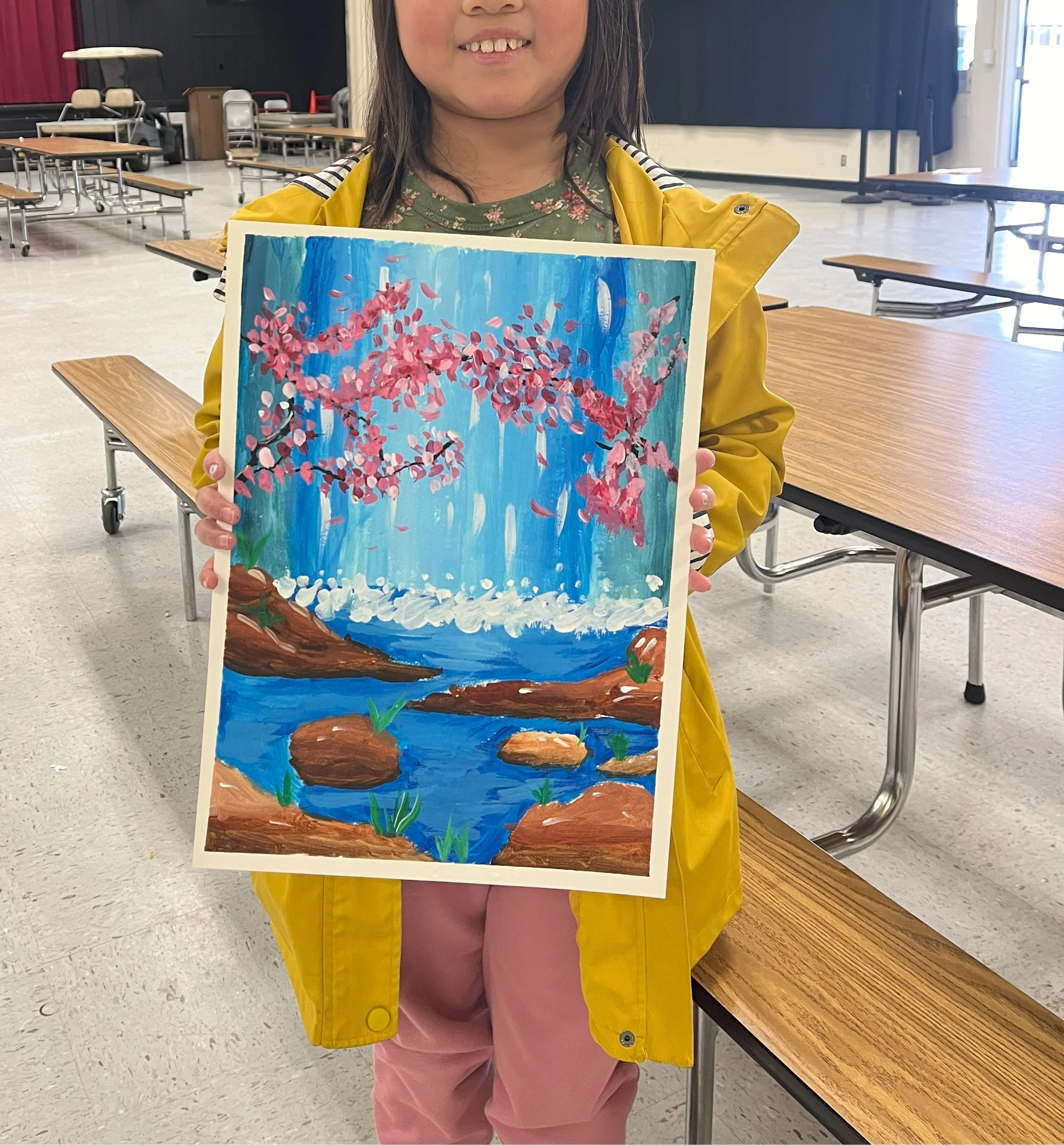 A child holding a painting depicting a springtime scene of cherry blossoms and a blue sky, in a large room with tables and chairs.