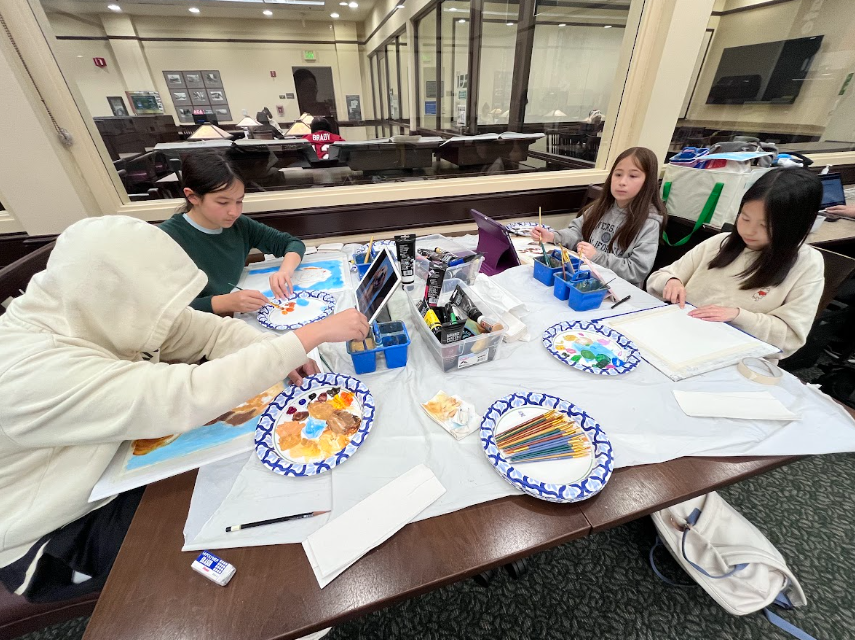 Four children sitting around a table engaging in arts and crafts activities, with art supplies and paper plates in front of them, in a room with large windows.