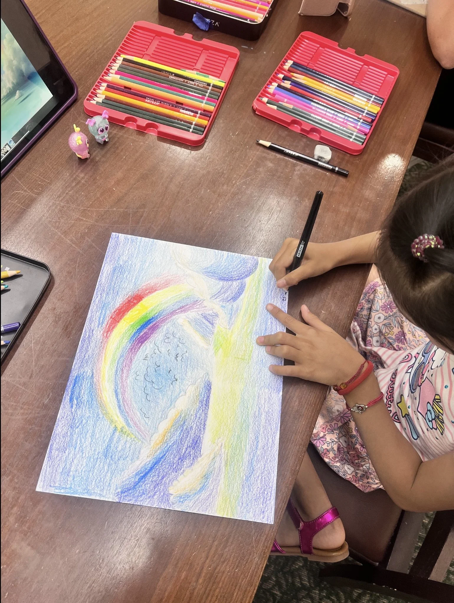 A girl coloring a rainbow with colored pencils on a sheet of paper on a wooden table. Two small toy creatures are beside her, and there are two red pencil cases filled with colored pencils. An iPad is visible on the left side of the image.