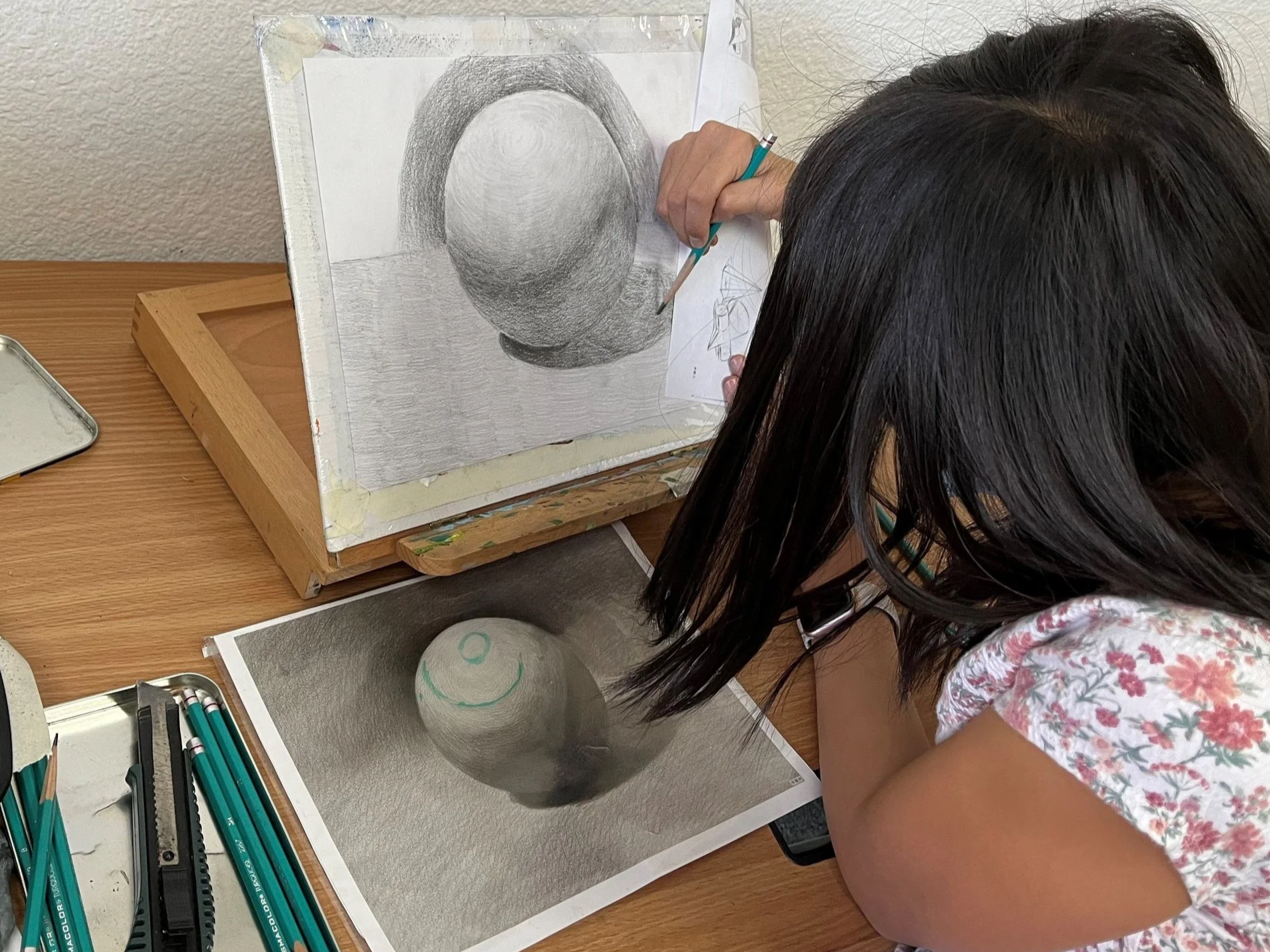 A girl drawing a sphere with pencil on paper, with a reference photo of a sphere smiling in front of her. Art supplies including colored pencils and a ruler are on the table.