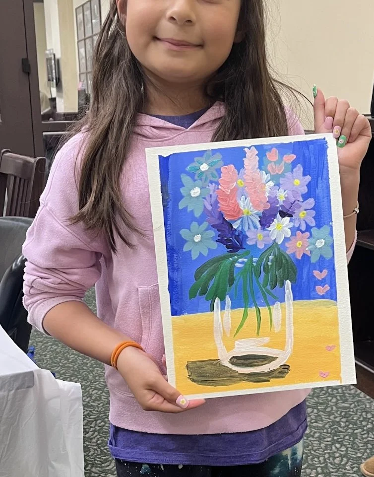 A young girl holding a colorful painting of a flower arrangement in a glass vase. The background is blue, and the table surface underneath the vase is yellow. The girl is smiling and wearing a pink hoodie.