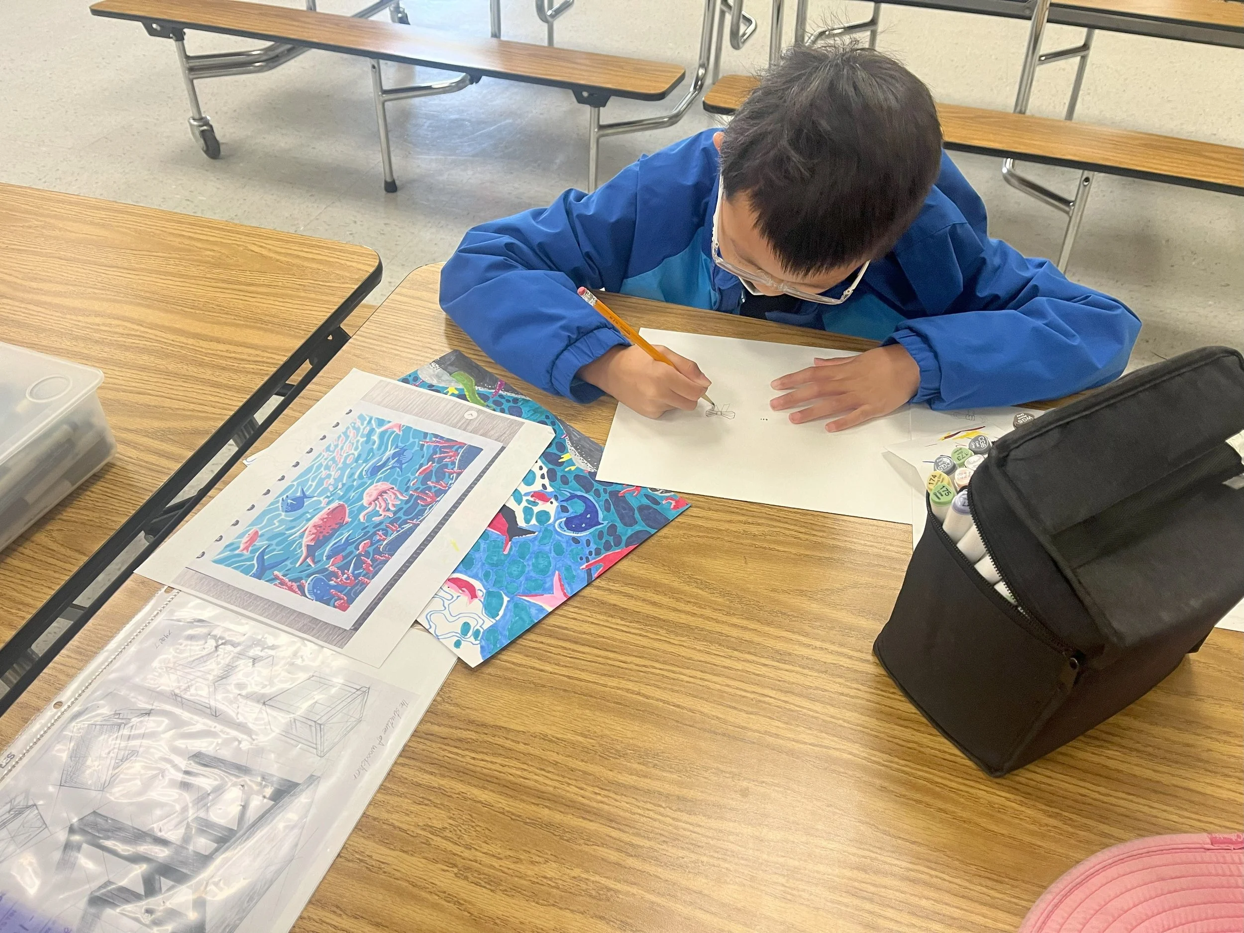 A student wearing glasses and a blue jacket drawing at a wooden desk with arts and crafts supplies, including printed color pages and a black bag nearby.