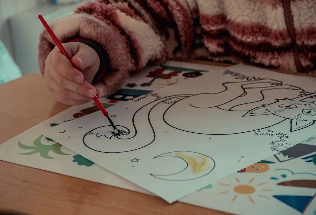Child coloring a unicorn drawing on a sheet of paper with a red marker, surrounded by other colorful papers.