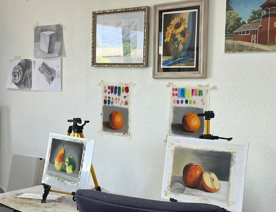 Art studio setup with still life paintings of fruits, drawing supplies, framed artwork, and sketches on the wall.