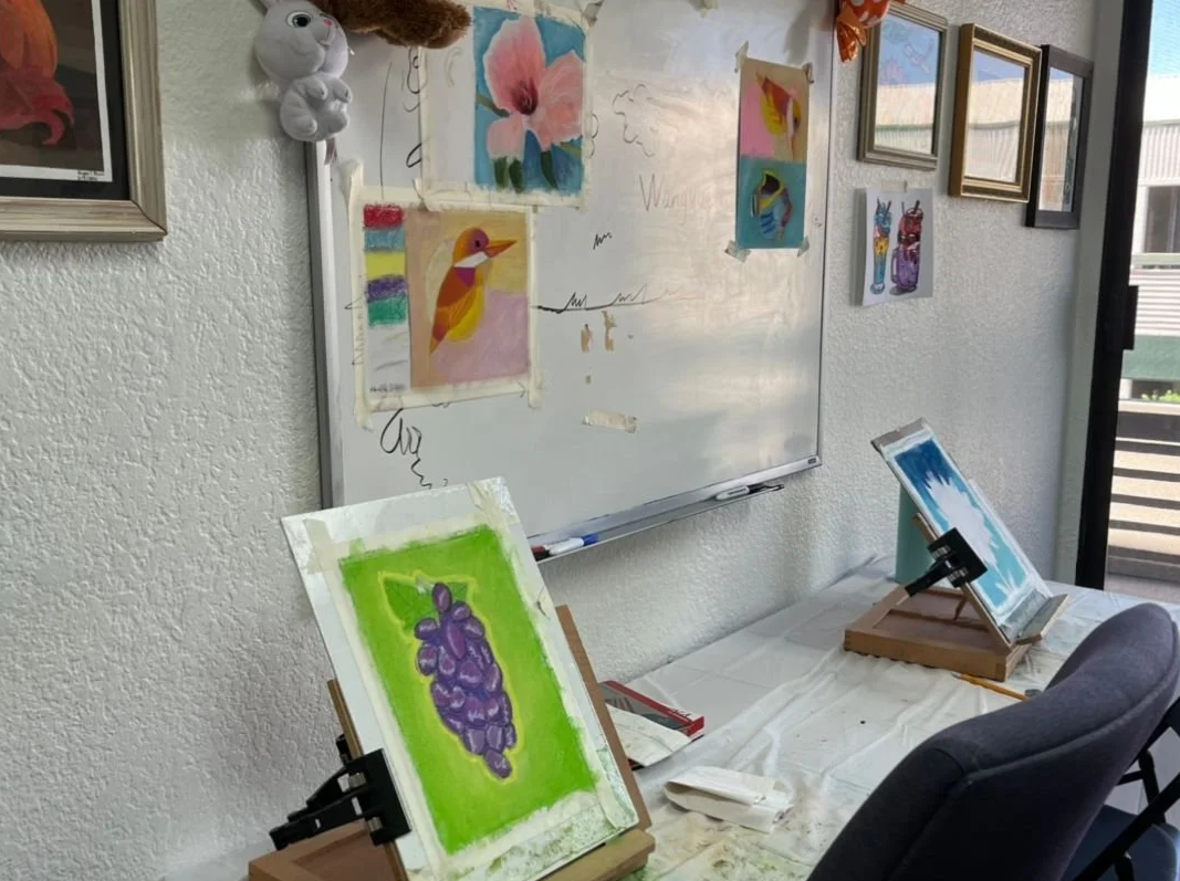 Art classroom with unfinished paintings on easels, a whiteboard with children's artwork, and framed pictures on the wall.