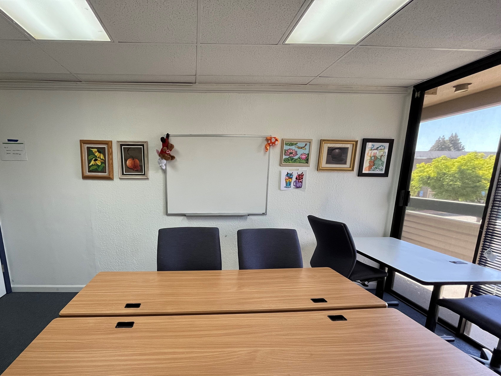 A meeting room with a whiteboard, framed artwork, and plush toys hanging on the wall. It has a wooden table, black office chairs, and an open window showing trees outside.