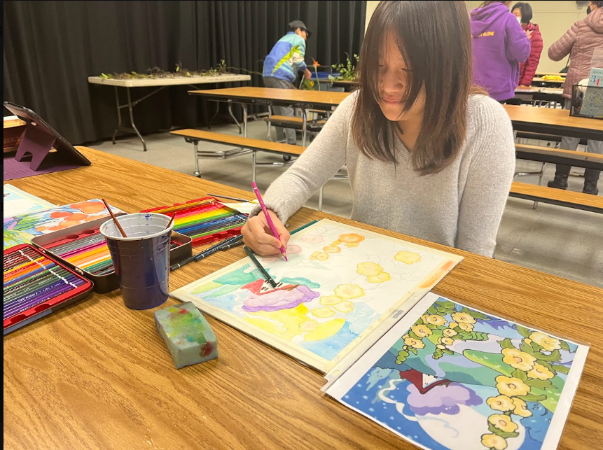 A woman sitting at a wooden table, painting with watercolors. She is surrounded by colorful painting supplies, with a watercolor painting of a tree with yellow flowers in front of her. Other people are working at tables in the background.