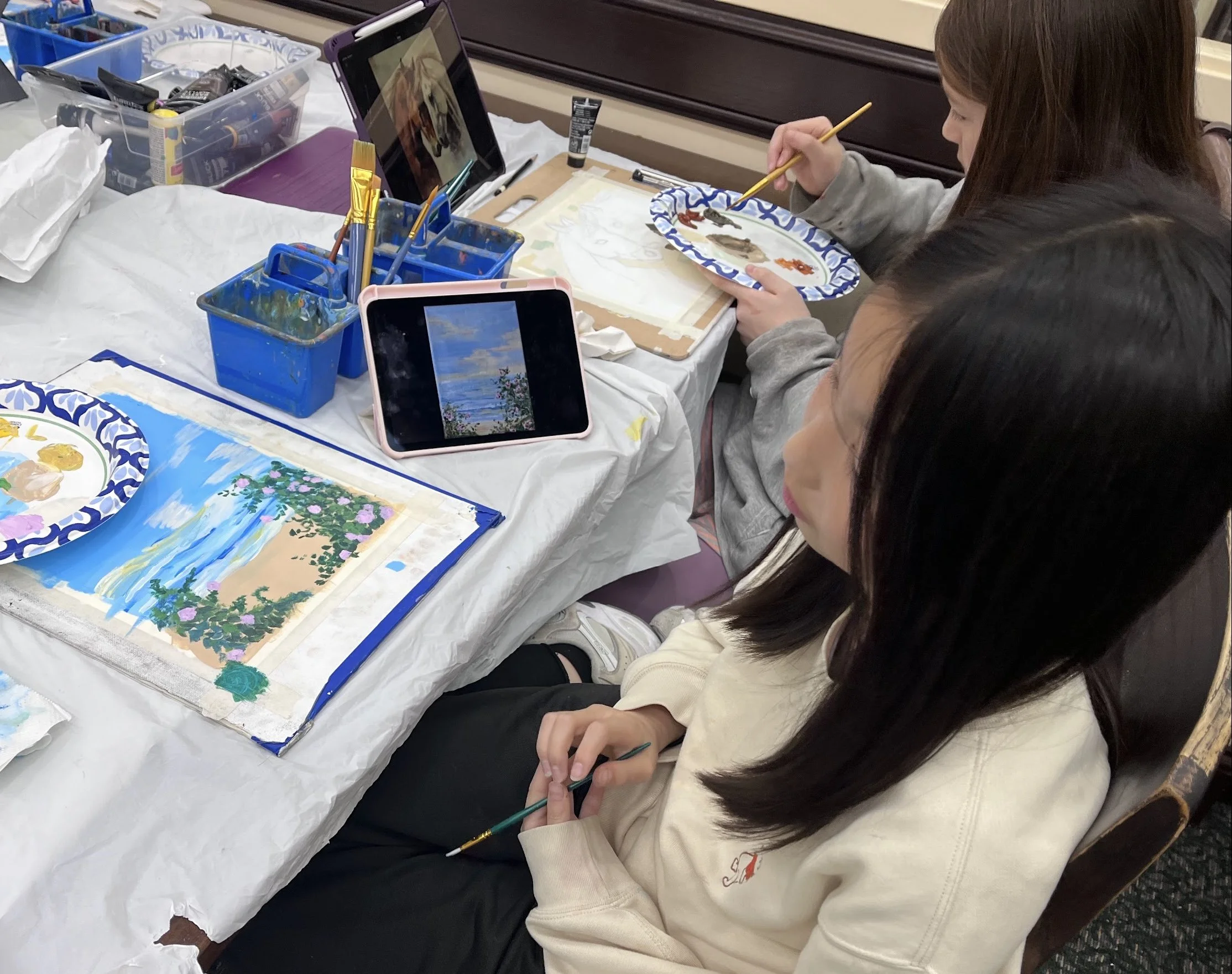 Several children sit around a table covered with a protective sheet, engaged in painting a landscape picture with trees, flowers, and a blue sky. They have various art supplies, such as paintbrushes and a palette, and are watching instructional videos on tablets.