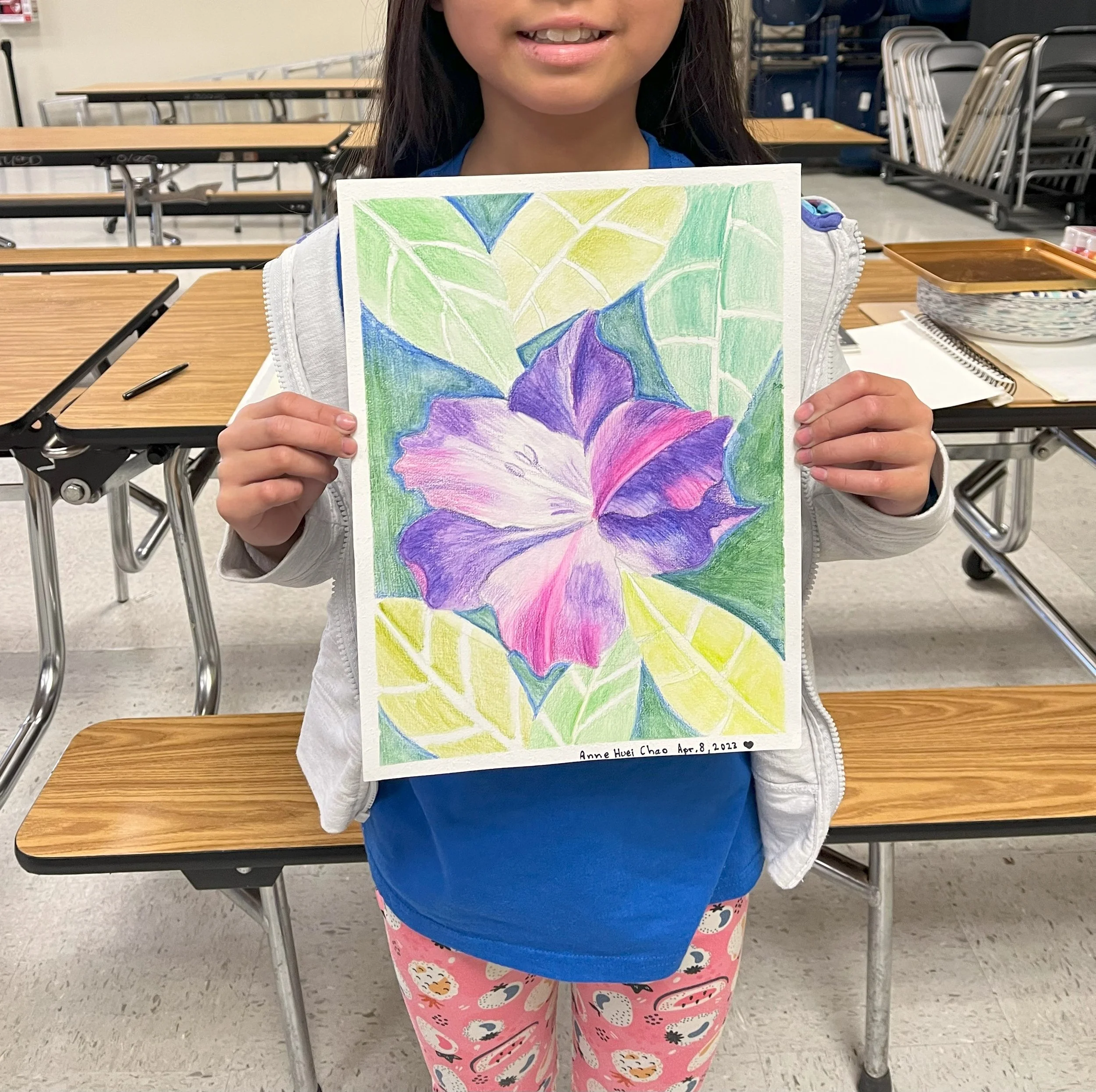 A young girl holding a colorful drawing of a flower with purple petals and green leaves in an indoor school setting.