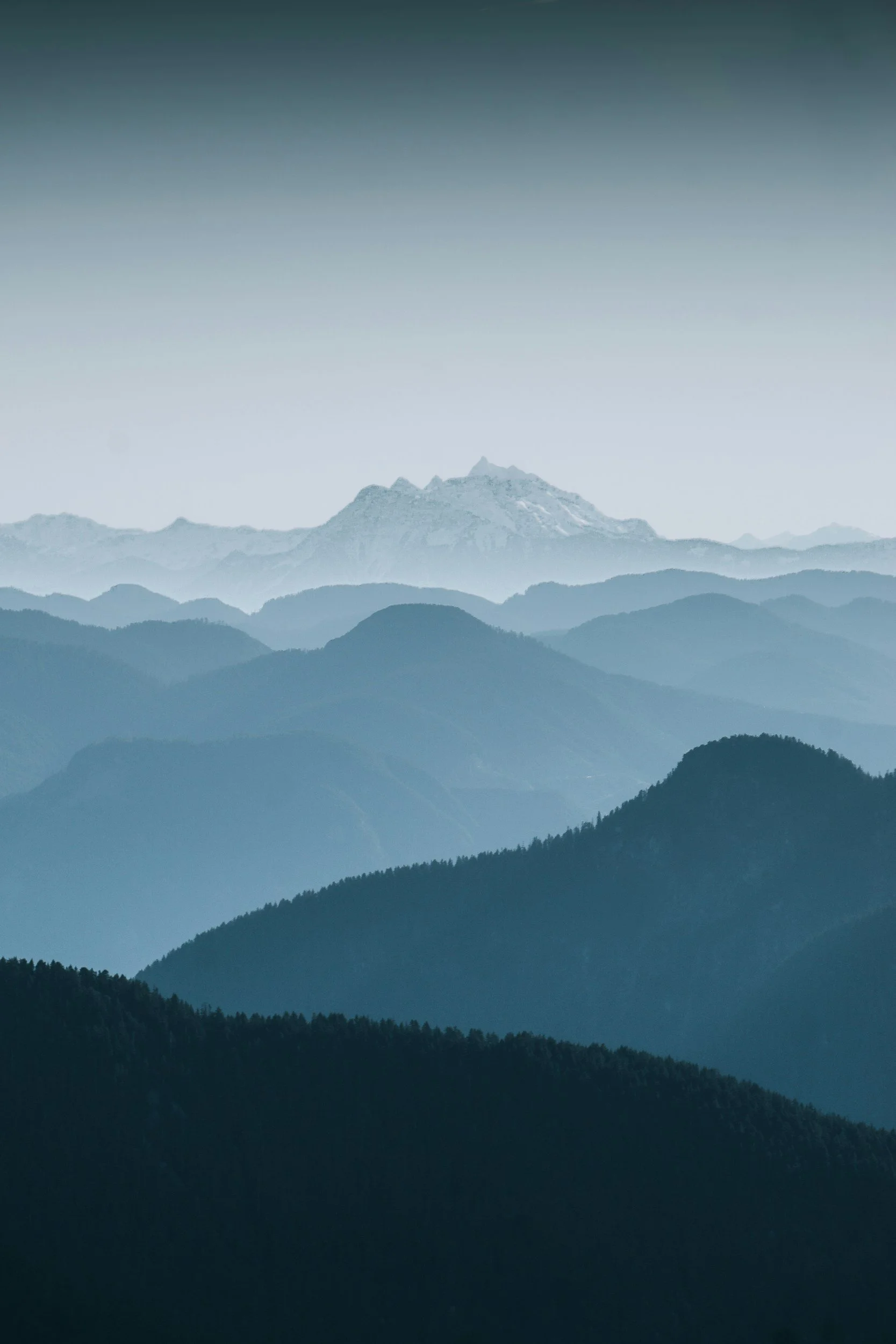 Layered mountain range with snow-capped peaks in the background, gradually darkening with distance, under a hazy sky.