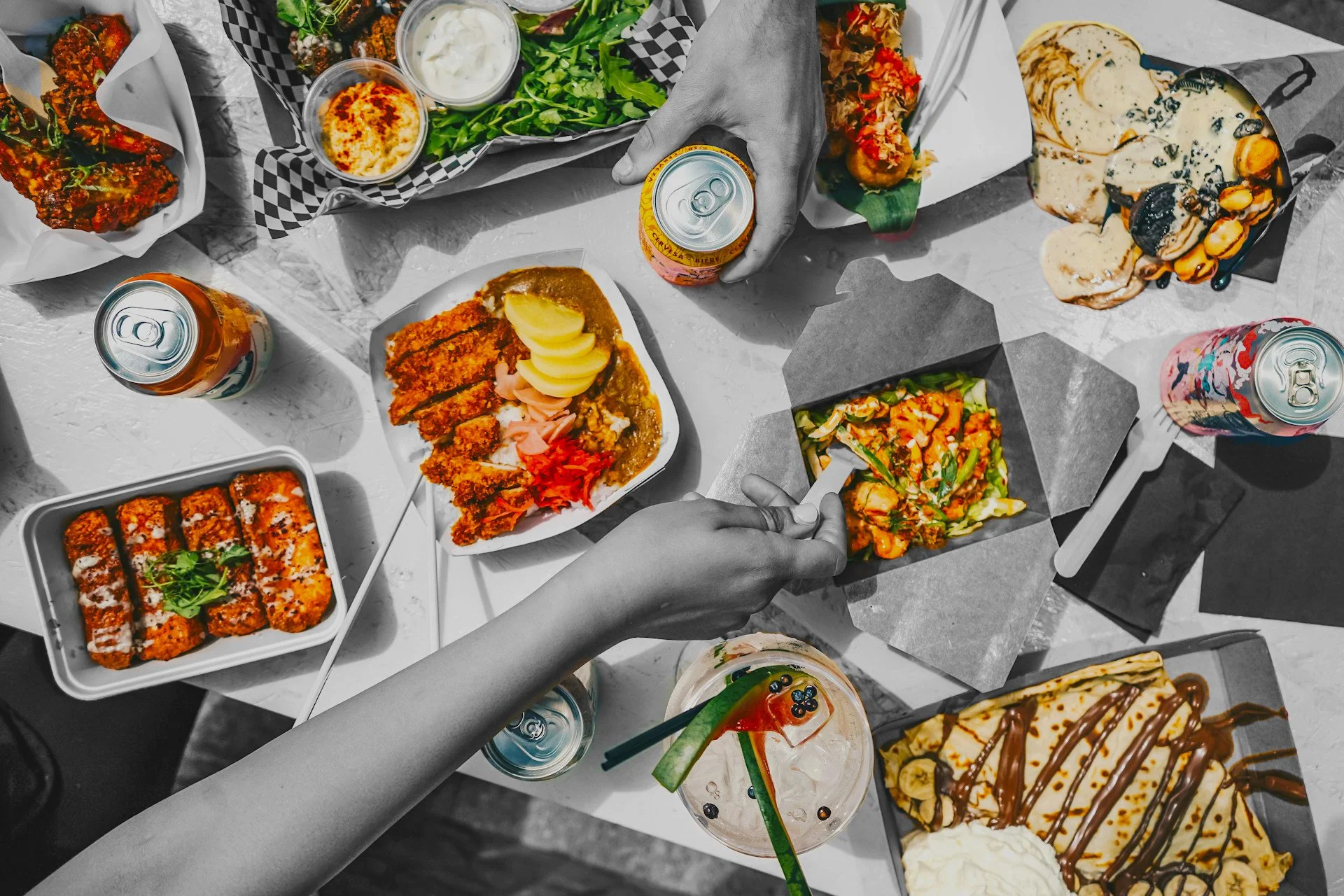 A table filled with assorted Asian and American dishes, including sushi, fried chicken, salad, ice cream, and drinks, with two people reaching for food.