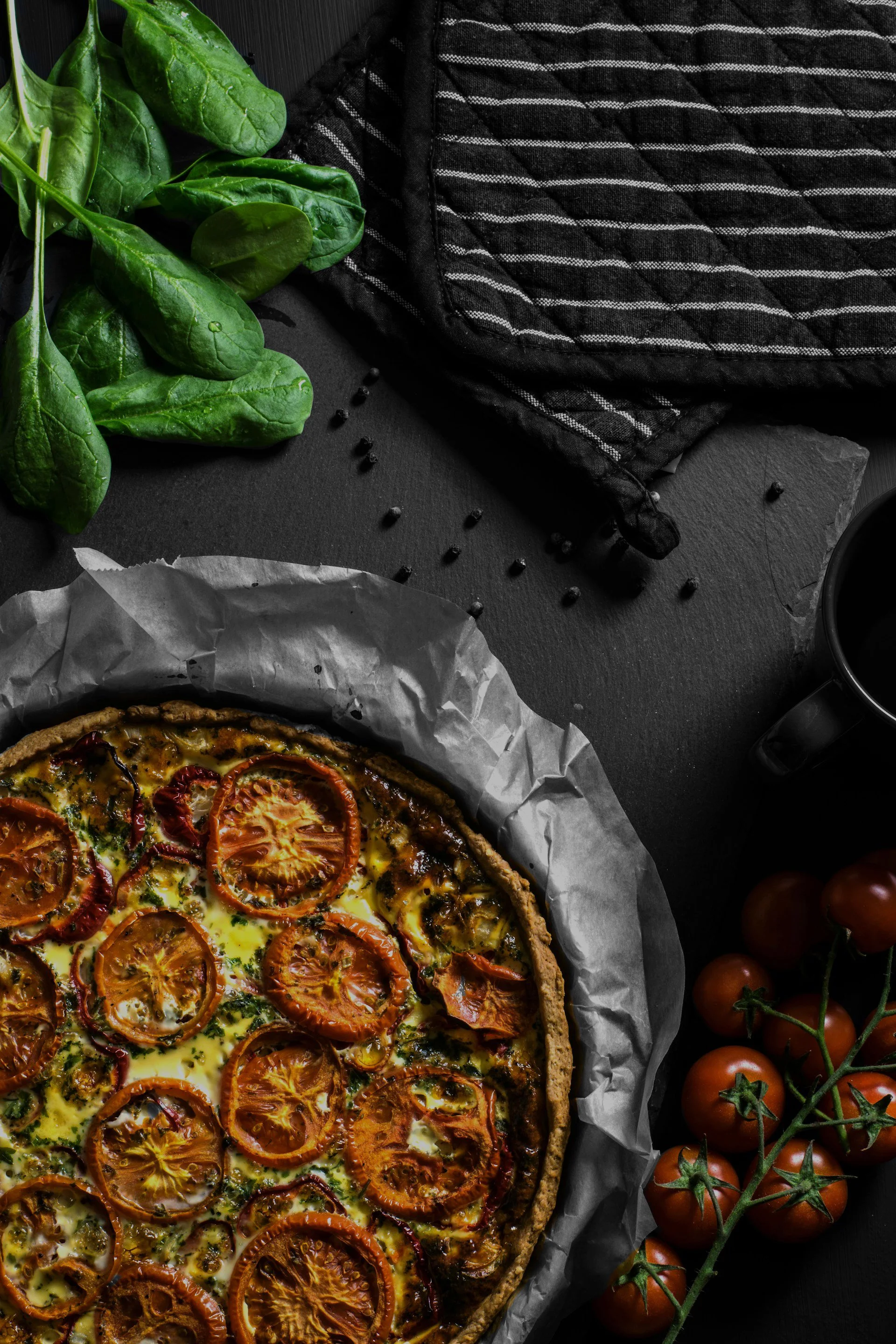 A freshly baked tomato and herb quiche with roasted tomatoes on top, against a dark background with a black and white cloth, green spinach leaves, cherry tomatoes, and scattered black peppercorns nearby.