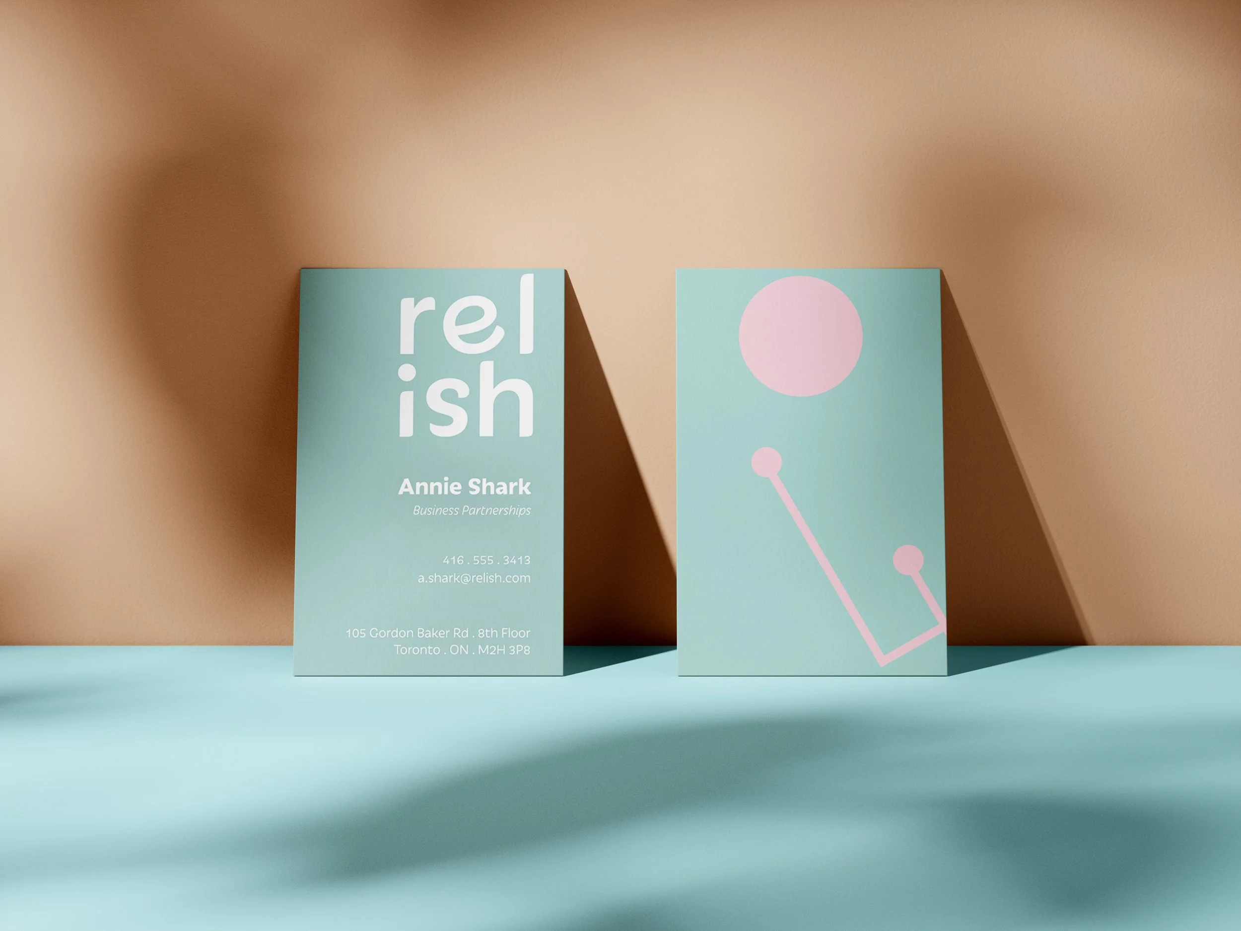 Business cards displayed on a turquoise surface. One card features the word 'relish' with the contact information of Annie Shark and her business details. The other card shows a pink dot with a line extending downwards, resembling a stylized question mark.