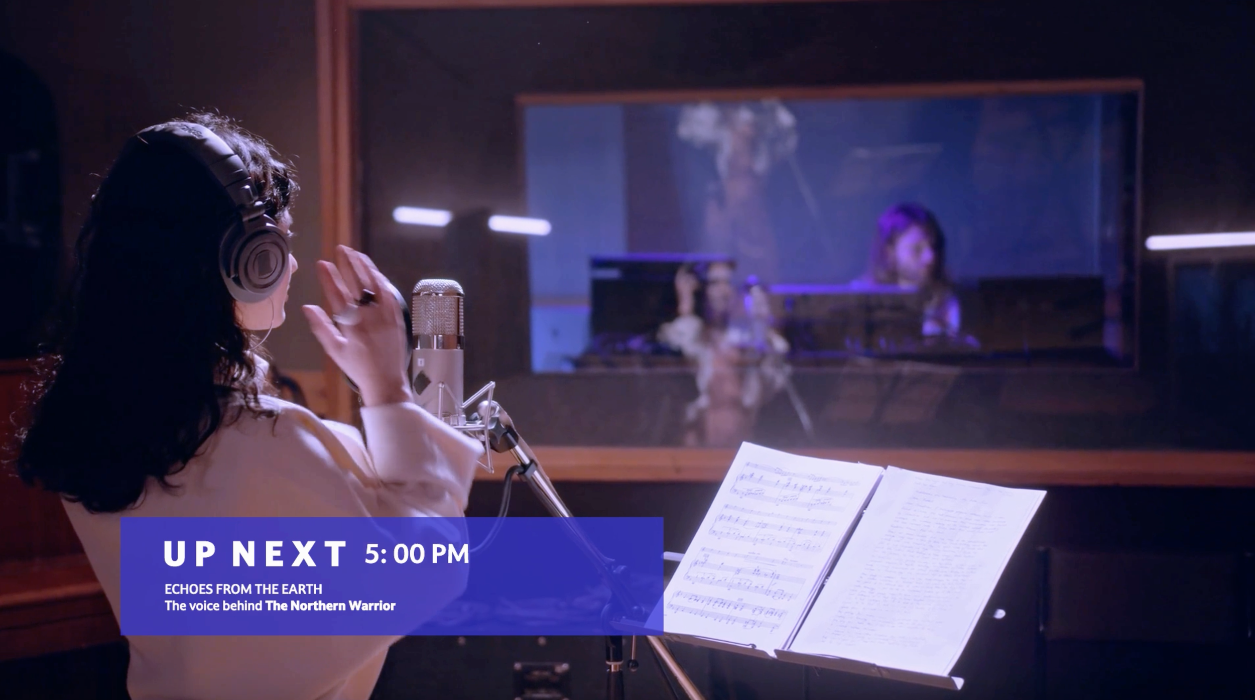 A woman in a recording studio is singing into a microphone while wearing headphones and reading sheet music. Behind a glass window, another person is visible in a different room, possibly the music producer. The lower part of the image features a blue overlay with white text that reads: 'UP NEXT 5:00 PM ECHOES FROM THE EARTH The voice behind The Northern Warrior.'