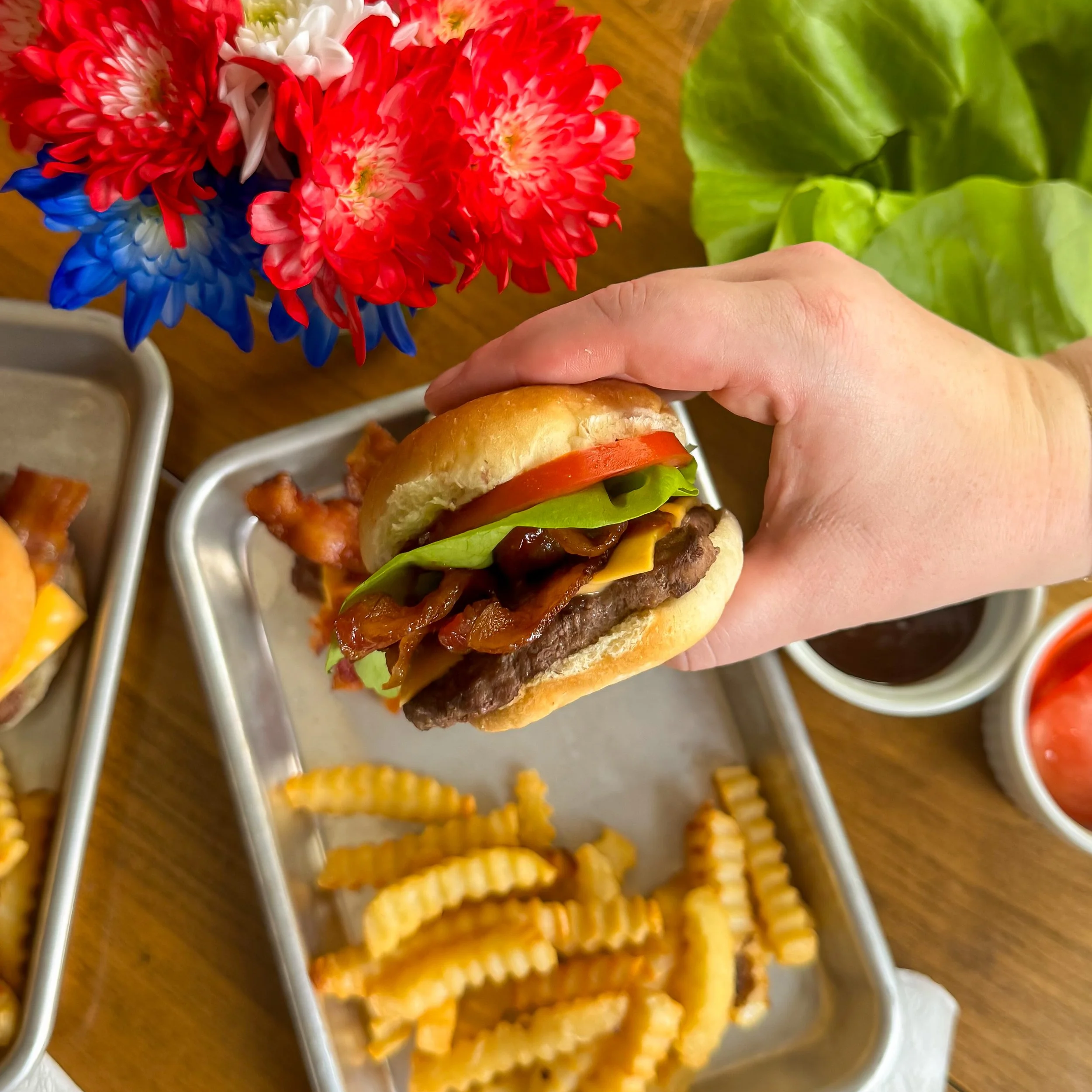 Close-up of colored flower bouquet with red, white, and blue flowers placed on a wooden surface near a hand holding a burger with lettuce, tomato, cheese, and bacon. Part of food trays and a sliced tomato in a bowl are visible nearby.