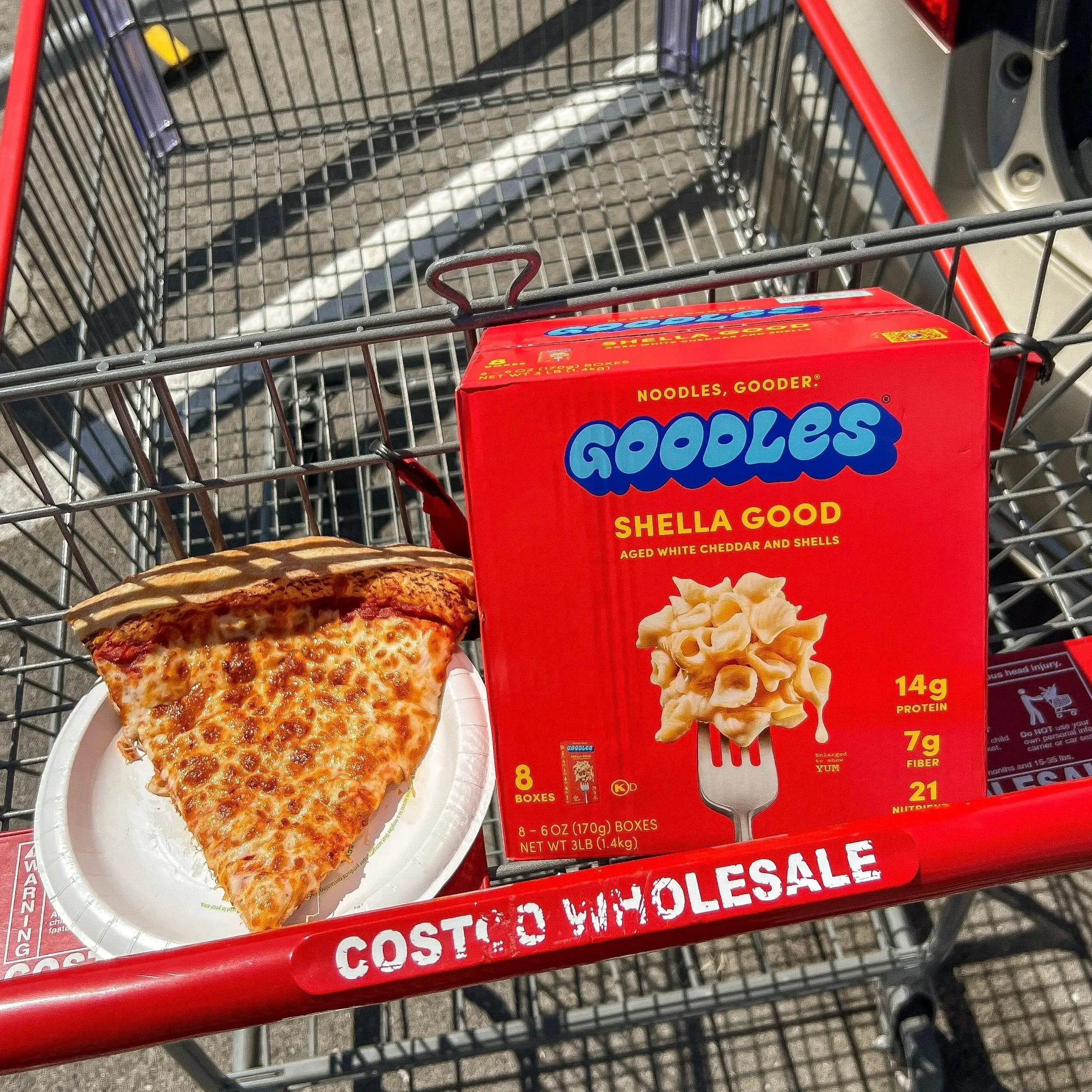 A shopping cart with a box of Goodles shell pasta and a slice of pizza on a paper plate. The cart is in a parking lot with red and black signage.