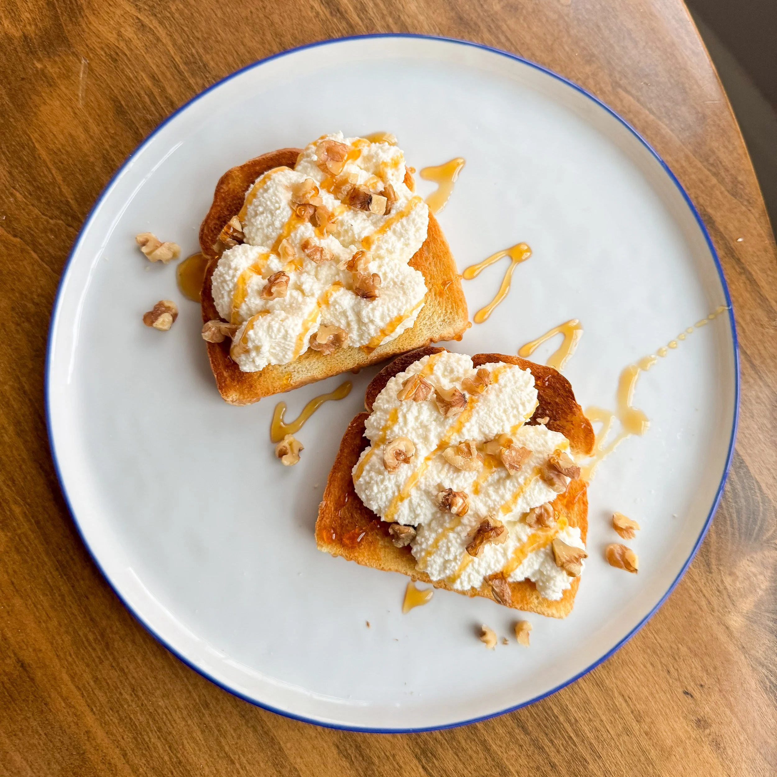 Slices of bread topped with whipped cheese, chopped nuts, and hoeny drizzle on a white plate with a blue rim on a wooden surface.