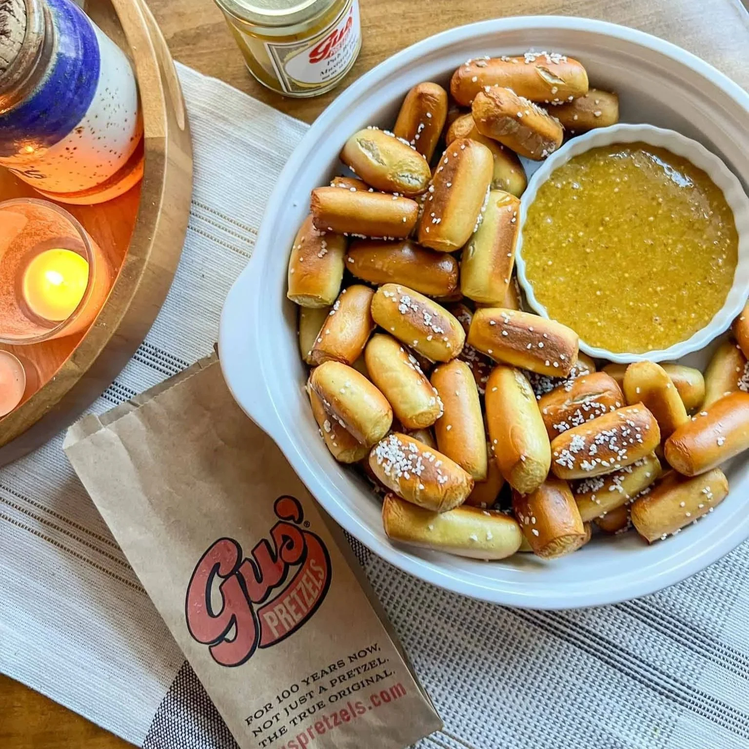 A plate of small pretzels with coarse salt, a bowl of mustard or cheese sauce, a glass jar of mustard, a lit candle in a glass holder, and a bottle of alcohol on a wooden table.