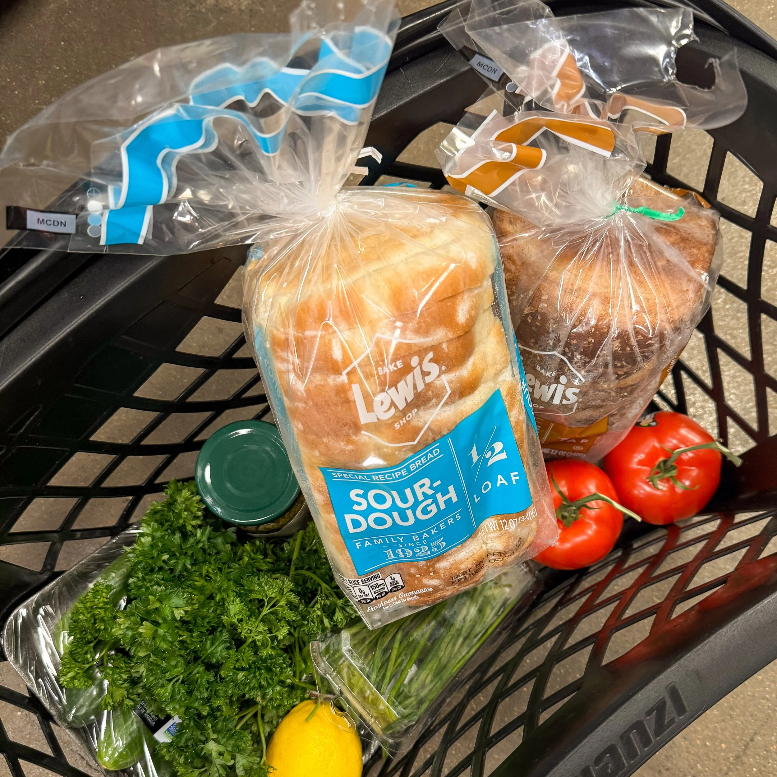 A grocery cart containing several plastic bags of bread, fresh vegetables including cherry tomatoes, lemons, parsley, and a green pepper, on a store floor.